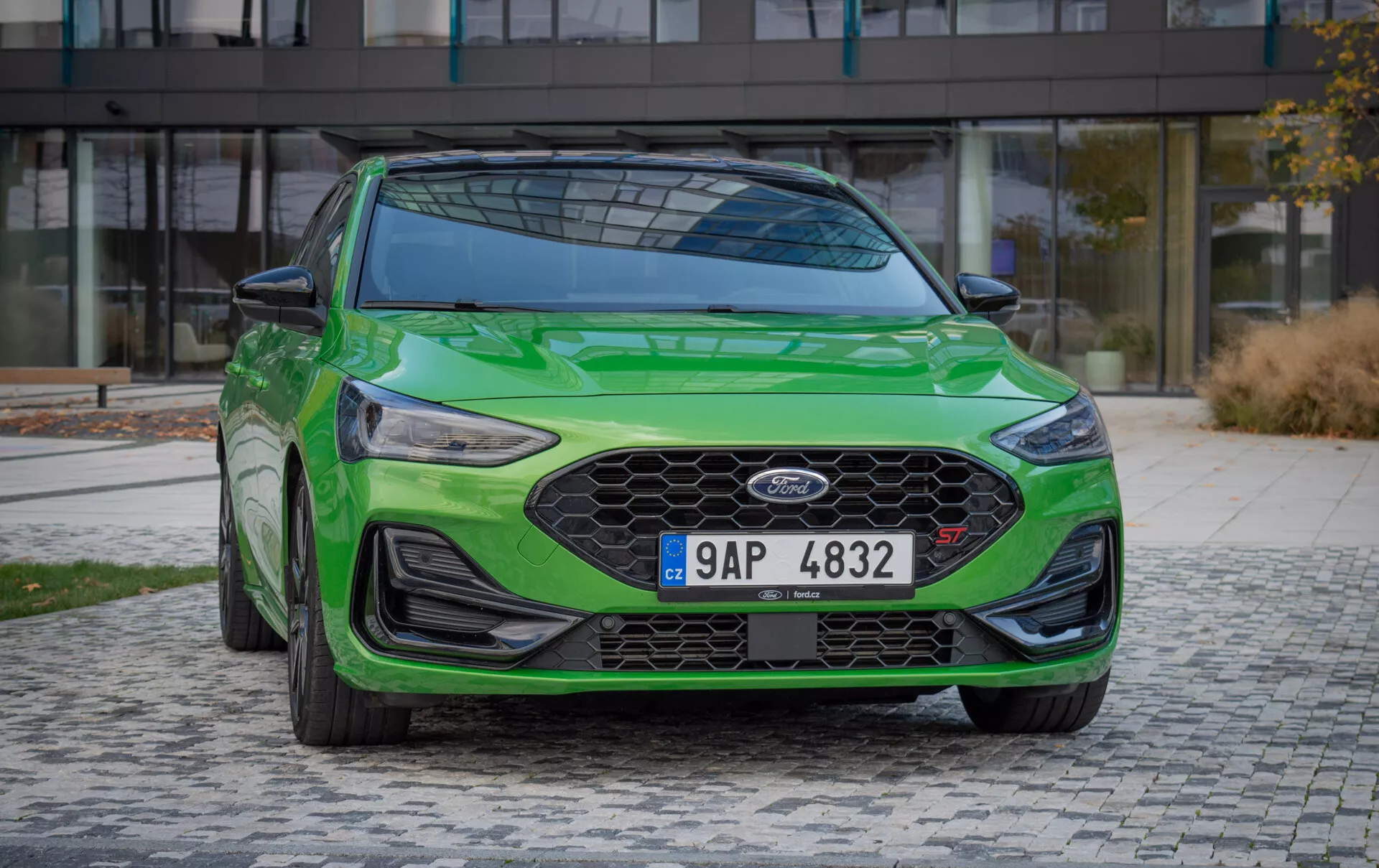 Ford Focus ST 2022 in verde vivace, mostrata da un angolo frontale sinistro, con una grande griglia a nido d'ape, fari LED eleganti, paraurti anteriore aerodinamico con spoiler, e ambientata su uno sfondo urbano moderno