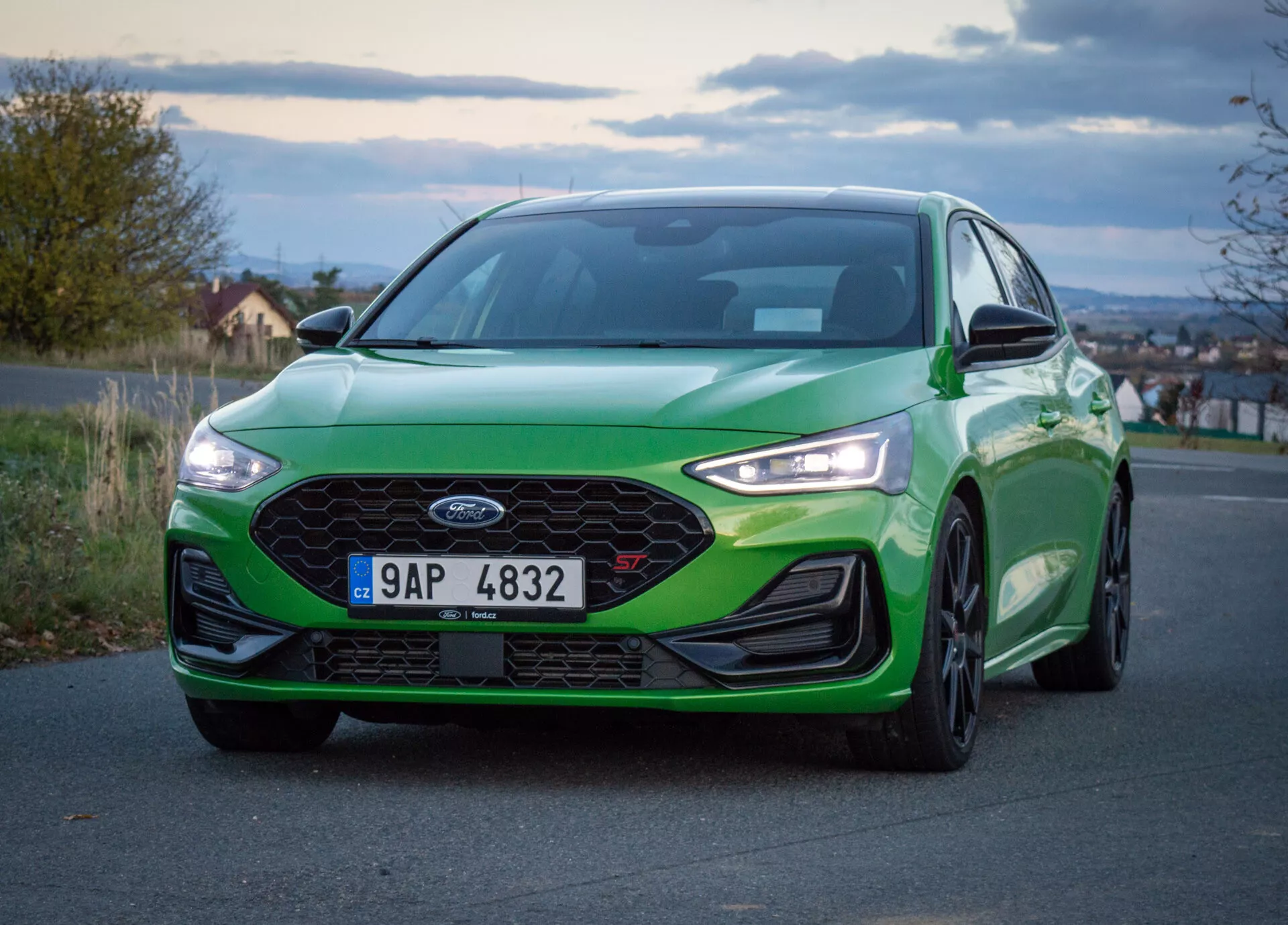 Ford Focus ST 2022, vista frontale angolata bassa, vernice verde con lucentezza vibrante, grande griglia a nido d'ape, fari LED e paraurti anteriore aerodinamico, su strada all'alba o al tramonto