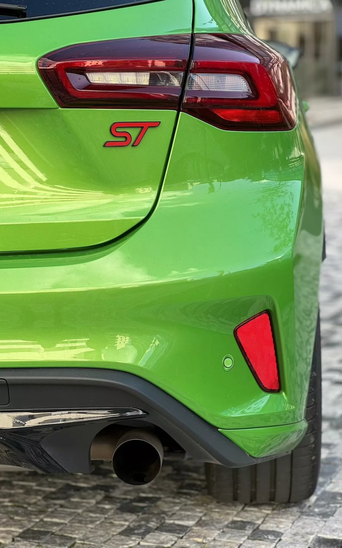 Ford Focus ST 2022 in verde brillante con badge ST, vista parziale di fanale posteriore rosso e trasparente, terminale di scarico cromato e elementi di design posteriore sportivo su superficie in ciottoli