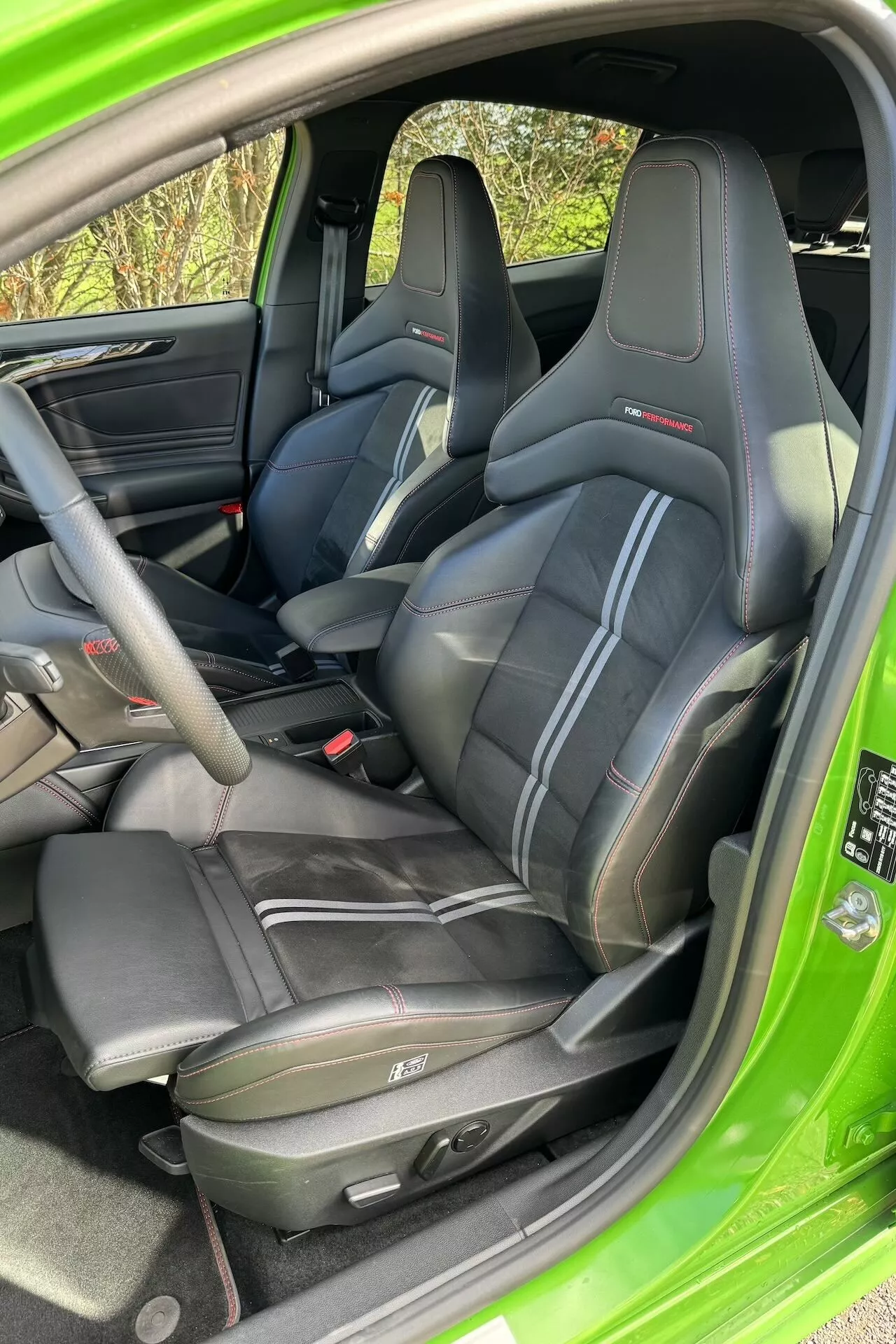 Interni del Ford Focus ST 2022 che mostrano sedili sportivi in pelle nera con cuciture a contrasto e poggiatesta integrati, design dettagliato di porta e console centrale, e luce naturale che illumina il vivace colore verde esterno