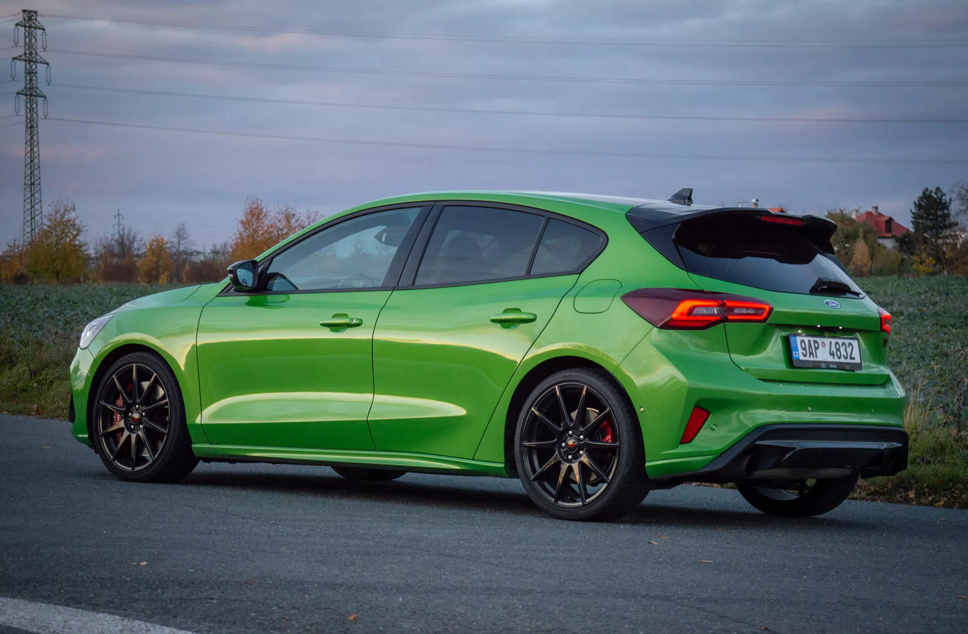 Ford Focus ST 2022 in verde lime con cerchi in lega sportivi e pinze dei freni rosse, catturata da un angolo posteriore laterale che mostra la sua sagoma hatchback, spoiler aerodinamico, terminali di scarico doppi integrati e luci posteriori LED in un tranquillo ambiente rurale al crepuscolo