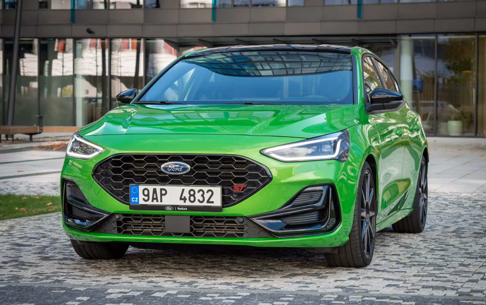 Ford Focus ST 2022 in verde vibrante, che mostra fari LED, griglia a nido d'ape e design aerodinamico da un angolo basso frontale dinamico a tre quarti con sfondo urbano moderno