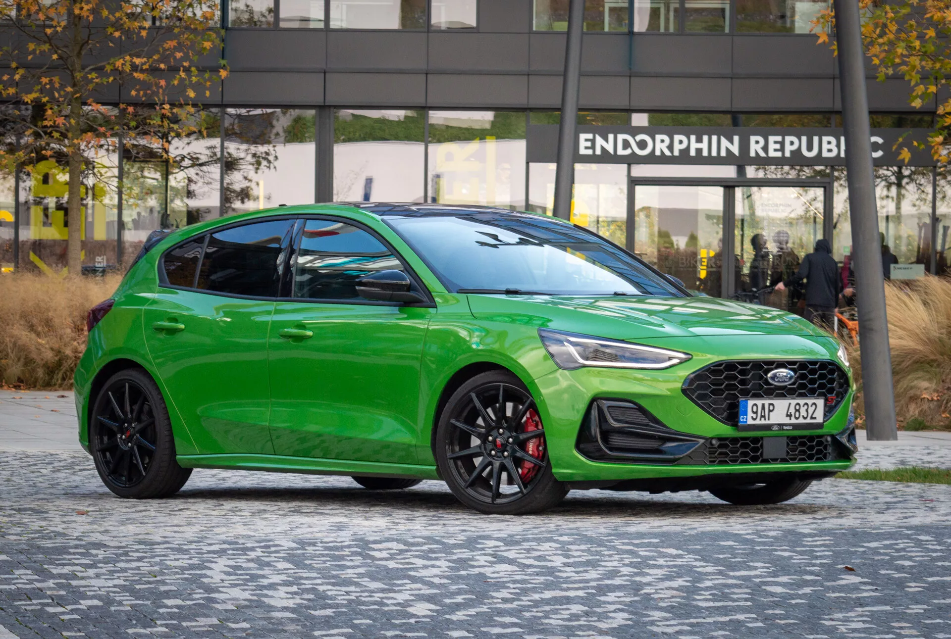 Ford Focus ST 2022 in verde brillante parcheggiata su pavimentazione decorata, mostra il suo profilo sportivo laterale-frontale, linee di design, dettagli visibili di fari e griglia, ruote orientate alle prestazioni e pinze dei freni, sotto un'illuminazione favorevole senza riflessi contro uno sfondo urbano con edificio per uffici moderno