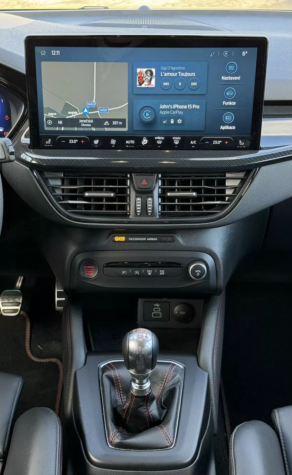 Interno Ford Focus ST 2022 con in primo piano la console centrale dotata di touchscreen per l'infotainment che mostra navigazione e connettività, controllo climatico bi-zona, leva del cambio manuale, comandi per la dinamica del veicolo e porta bicchieri, evidenziando tecnologia e dettagli di design premium.