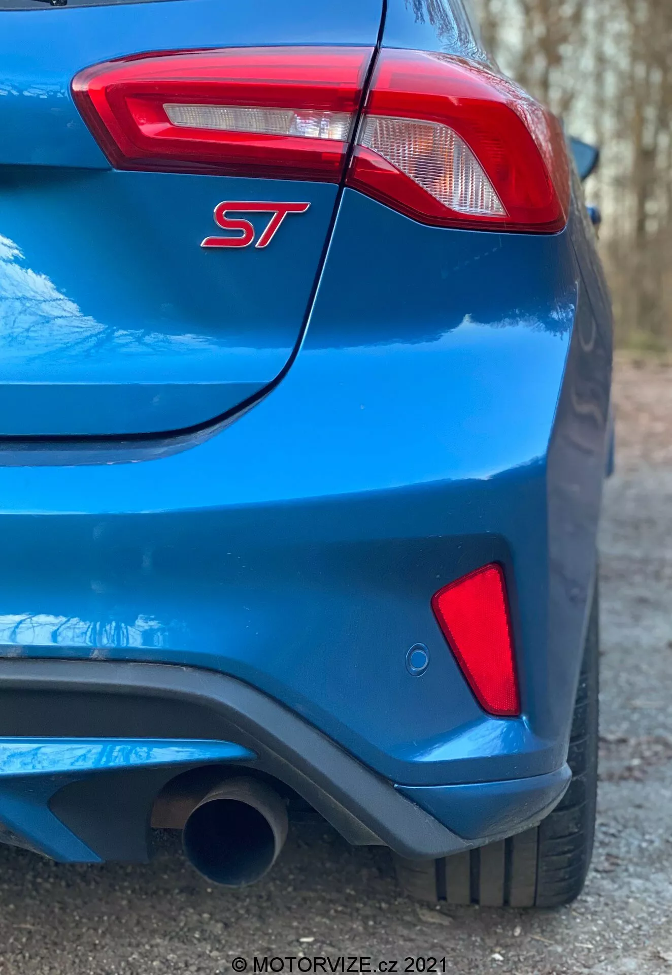 Vista ravvicinata e angolata della parte posteriore di una Ford Focus ST 2022 blu. Il quadrante inferiore destro dell'immagine evidenzia un modello orientato alle prestazioni indicato dal distintivo 'ST'. Le caratteristiche notevoli includono un singolo tubo di scarico prominente nella parte inferiore sinistra, un paraurti posteriore scolpito con un design integrato tipo diffusore e una luce posteriore moderna e parzialmente visibile con elementi LED. La vernice blu vibrante ha una finitura lucida e una piega sottile corre sopra la luce posteriore lungo il portellone, migliorando l'estetica sportiva. L'angolo della foto cattura l'assetto e l'altezza dell'auto, focalizzandosi sulla trama e sui dettagli del design del paraurti posteriore.