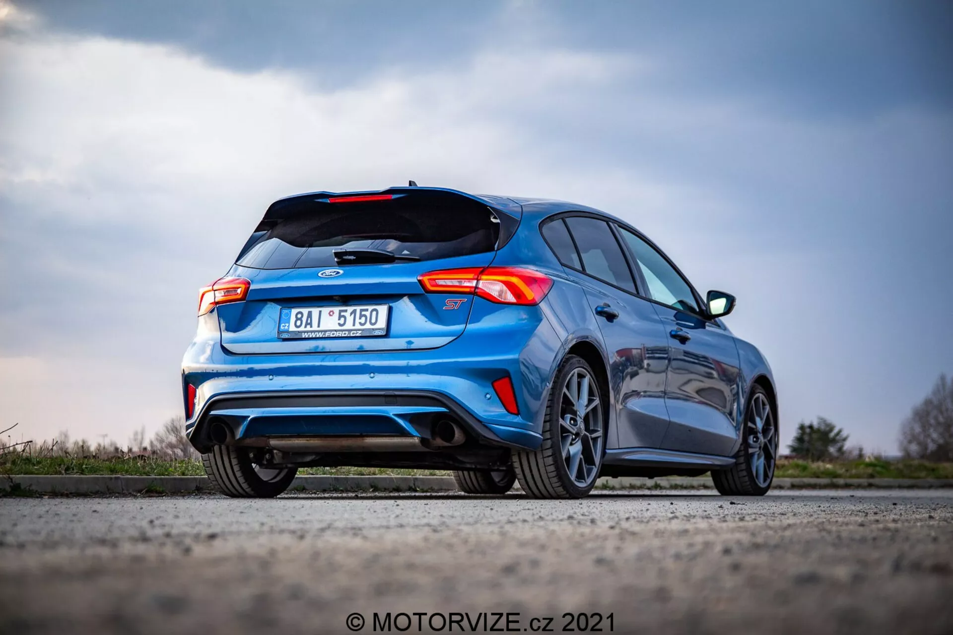 Immagine della Ford Focus 2022 da un'angolazione posteriore bassa, che mostra il design hatchback, il logo del produttore sul bagagliaio, l'esclusivo design delle luci posteriori, paraurti posteriore con diffusore, doppi terminali di scarico, spoiler posteriore, cerchi in lega, targa centrata, colore blu vibrante, contro uno sfondo di paesaggio naturale sfocato e cielo. L'auto è ferma su una superficie piana, catturata in nitido fuoco con effetto bokeh sullo sfondo.