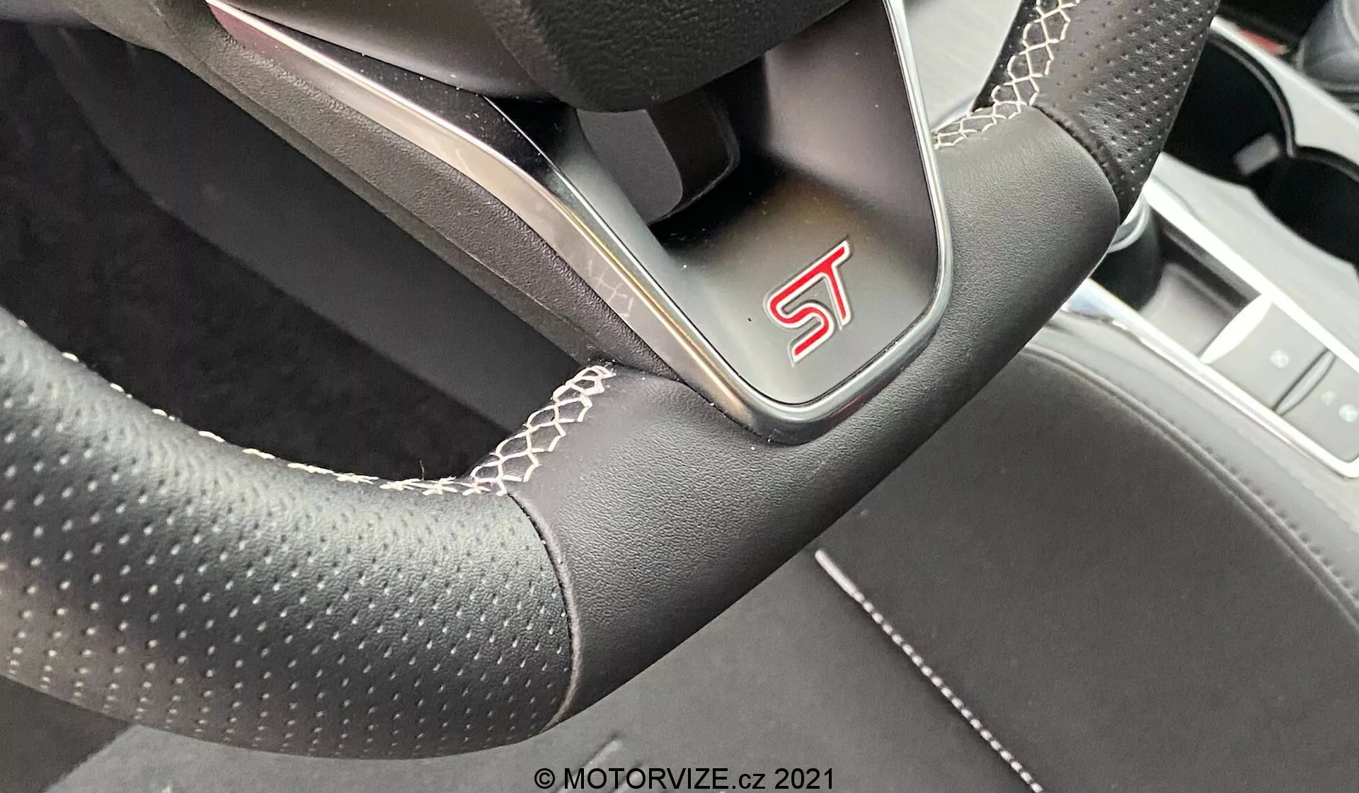 Vista ravvicinata di un volante moderno e sportivo dalla prospettiva del conducente in una Ford Focus 2022, caratterizzato da un emblema 'ST', materiale in pelle traforata, cuciture bianche, design leggermente appiattito sul fondo e raggi metallici che indicano una lavorazione orientata alle prestazioni.