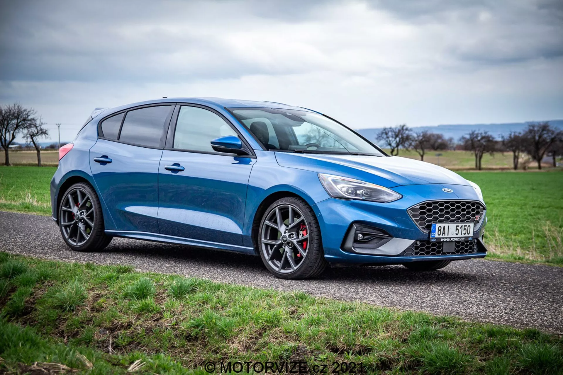 Ford Focus 2022 blu hatchback catturato da un'angolazione laterale in un'ambientazione naturale lungo una strada di campagna. Il veicolo presenta un moderno design dei fari a LED, una griglia frontale prominente, fendinebbia sul paraurti inferiore, cerchi in lega scuri con pneumatici a basso profilo e pinze dei freni rosse. Il profilo laterale rivela linee eleganti della carrozzeria, specchietti laterali pieghevoli, maniglie delle portiere, finestrini leggermente oscurati e tappo del carburante, il tutto contribuendo alla sua estetica sportiva.