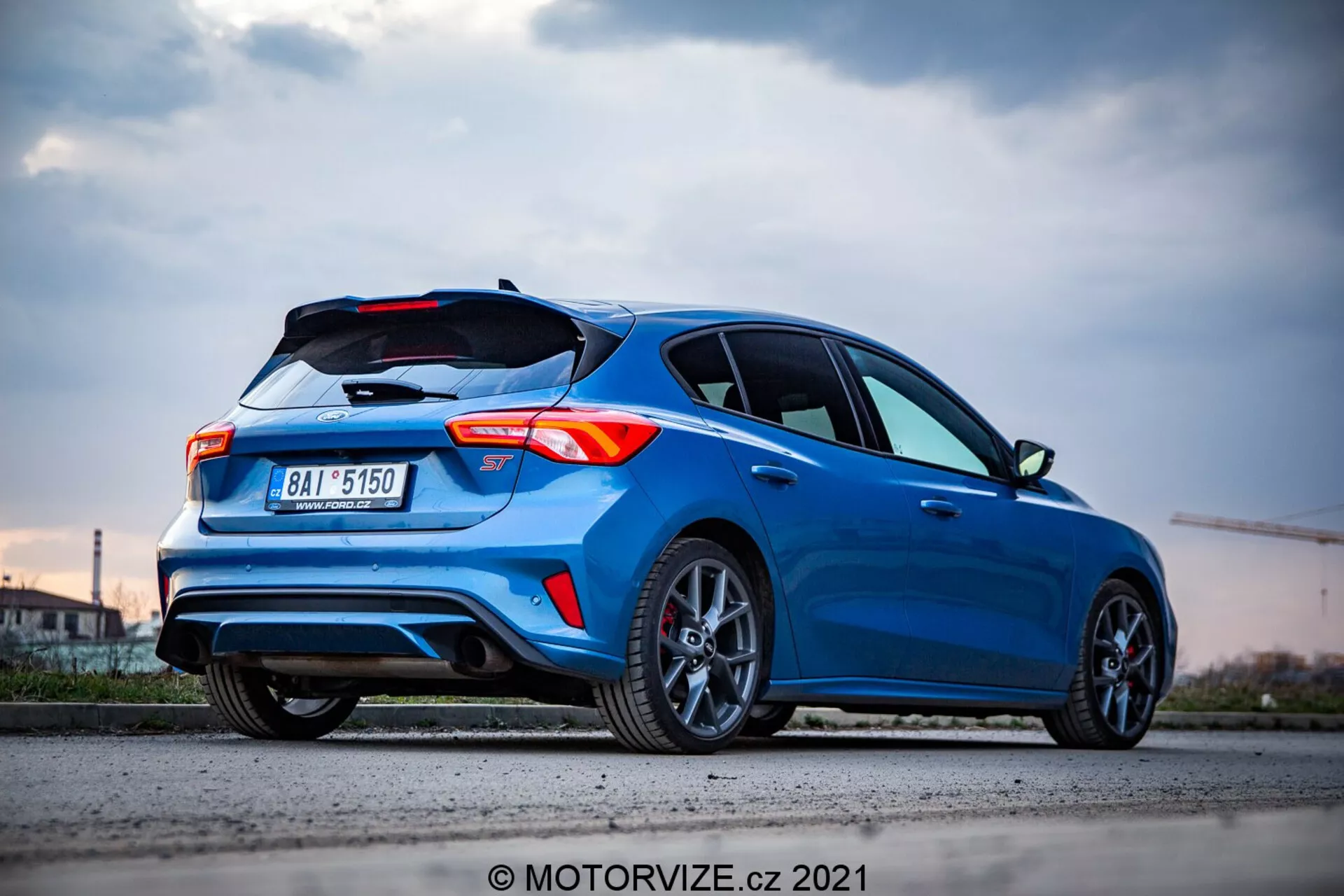 Ford Focus 2022 hatchback blu vibrante con caratteristiche di design sportivo come spoiler sul tetto, elementi aerodinamici e linee affilate, visto da un basso angolo posteriore a tre quarti. L'auto mostra fari posteriori a LED moderni, doppio scarico, cerchi in lega multiraggio scuro e un design del paraurti dinamico. Fotografiato al crepuscolo, enfatizzando l'esterno elegante dell'auto e lo sfondo urbano, con riflessi di superficie che indicano un veicolo pulito e ben tenuto.
