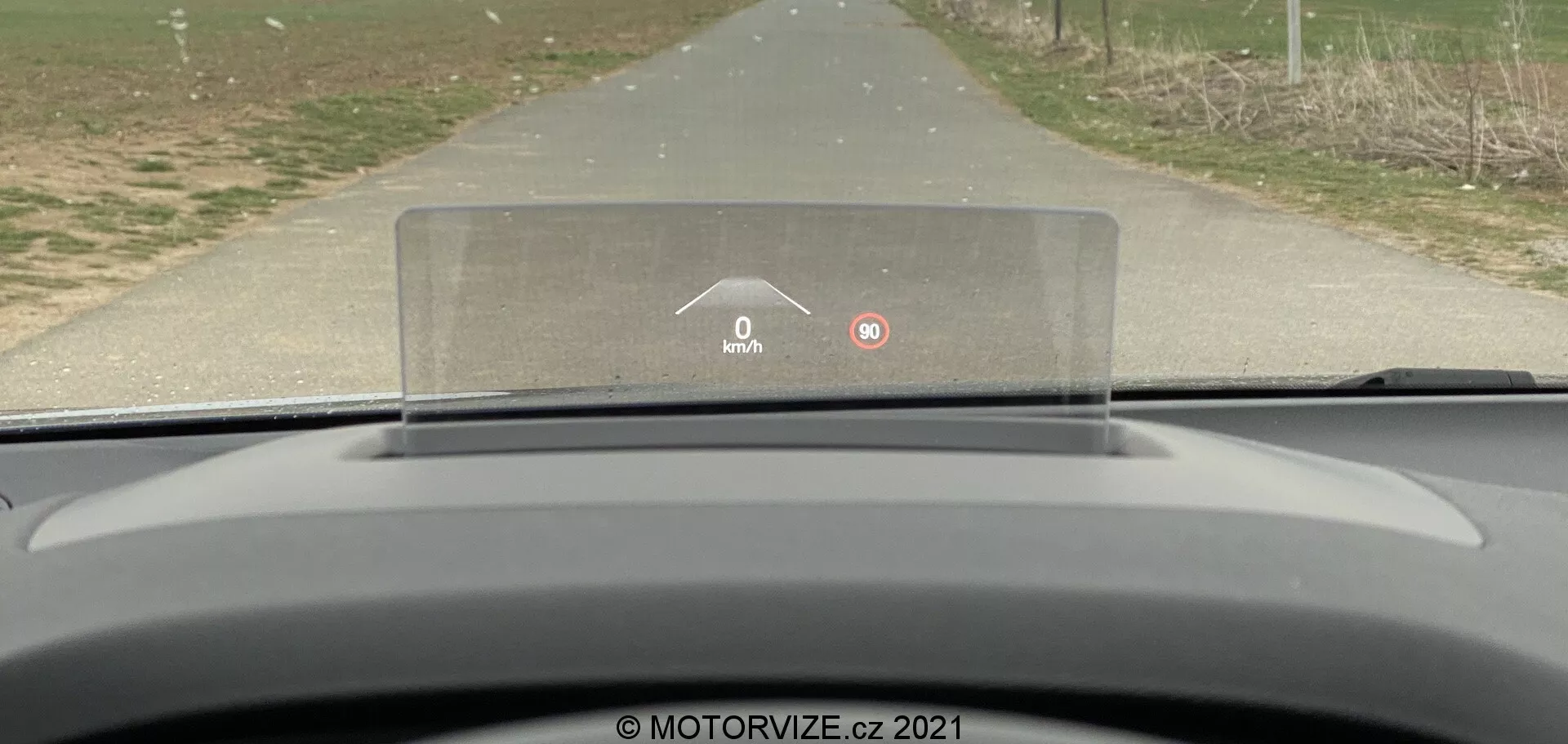 Vista dal sedile del conducente di una Ford Focus 2022, che mostra il cruscotto curvo e minimalista e il display head-up (HUD) prominente proiettato sopra il volante. L'HUD mostra una velocità di 0 km/h e un'icona di avviso, suggerendo che qualcosa richiede l'attenzione del conducente. L'auto sembra essere ferma su una strada rurale o suburbana, con erba e alberi ai lati della strada, enfatizzando un ambiente tranquillo e non urbano.