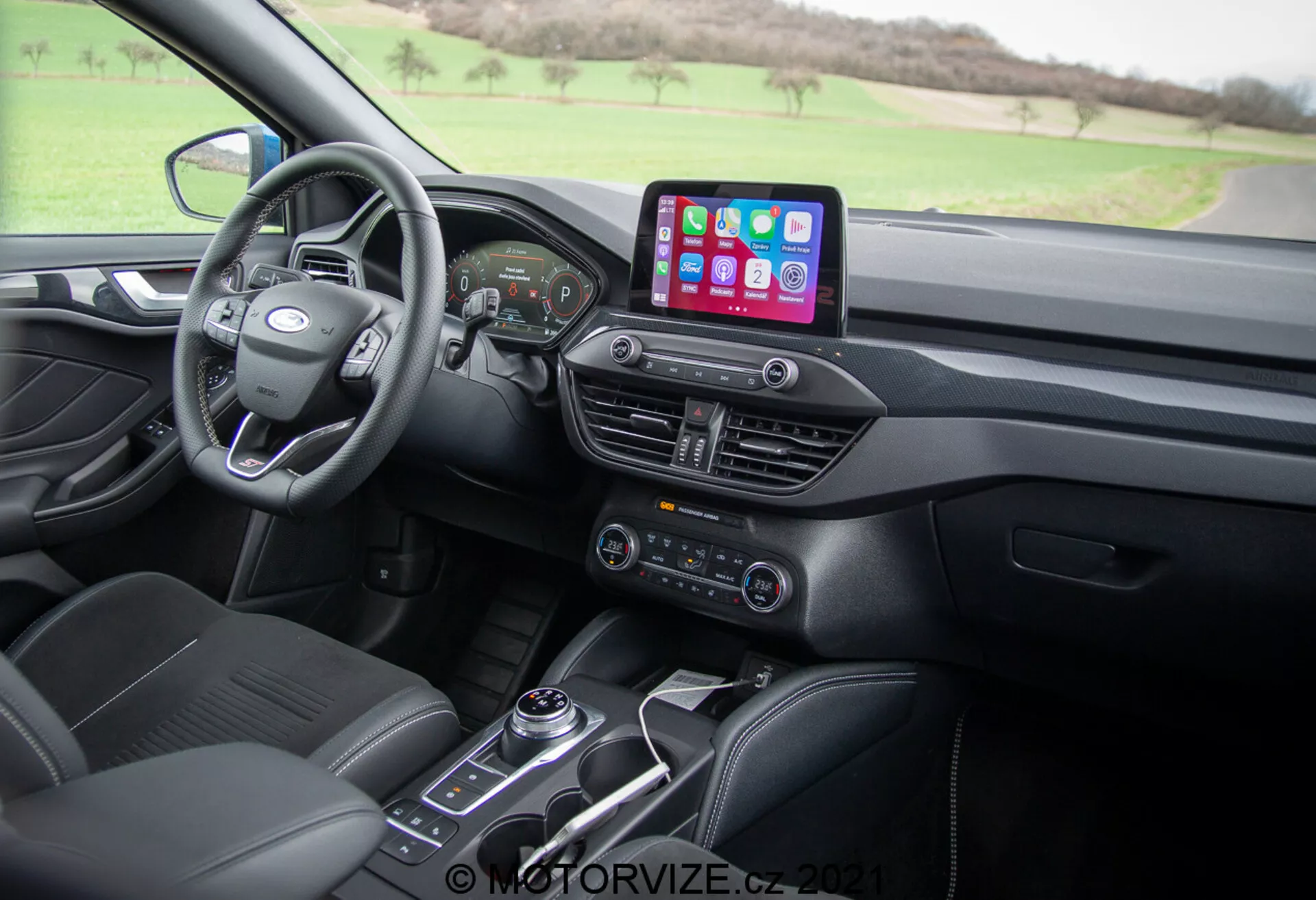 Vista dell'abitacolo del Ford Focus 2022 dal sedile del passeggero anteriore, che mostra il volante multifunzione moderno, cruscotto digitale, sistema di infotainment con connettività per smartphone come Apple CarPlay e Android Auto, sistema di controllo del clima con pulsanti per i sedili riscaldati, leva del cambio automatico e sedili in pelle con motivi di cucitura.