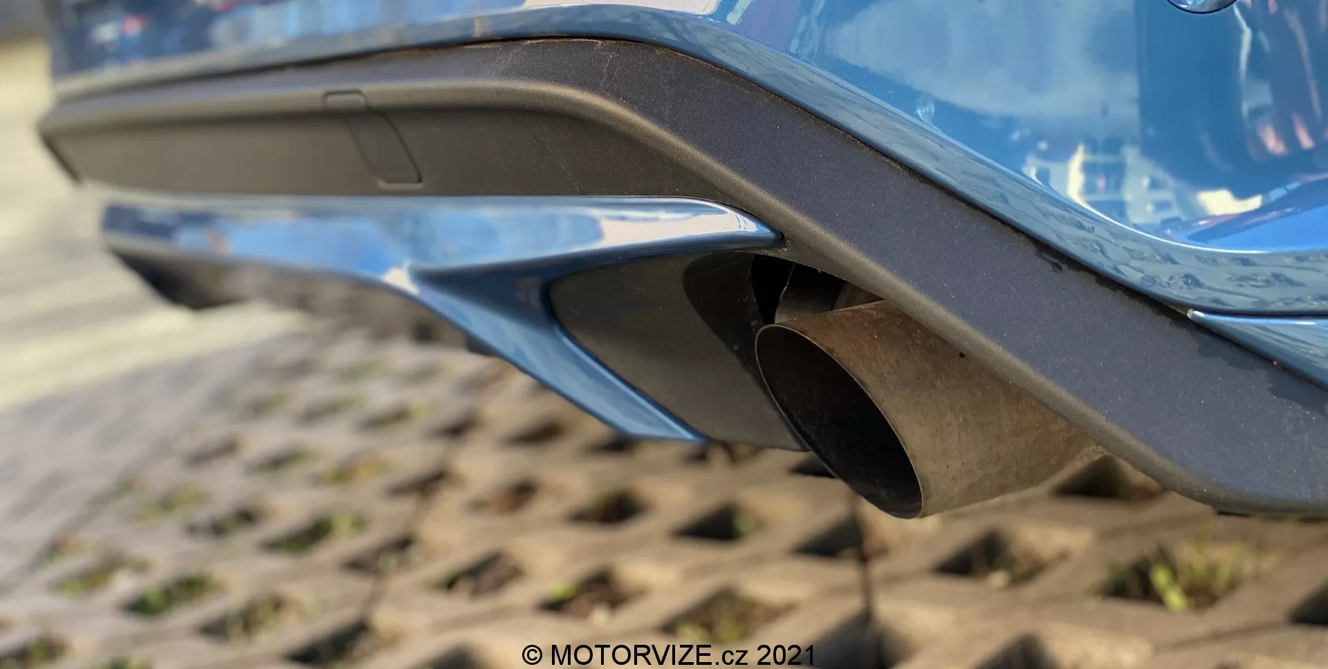 Immagine ravvicinata della sezione posteriore di una Ford Focus 2022, con particolare attenzione al sistema di scarico. L’immagine, scattata da un’angolazione bassa, mette in risalto la parte inferiore del paraurti posteriore con un design moderno e aerodinamico, vernice lucida e caratteristiche integrate come prese d'aria o diffusori. Al centro, un terminale di scarico circolare in metallo con una finitura leggermente consumata sporge dal paraurti. La superficie testurizzata sotto suggerisce un’area di parcheggio industriale o di uso intenso, con un'occhiata al telaio del veicolo visibile.