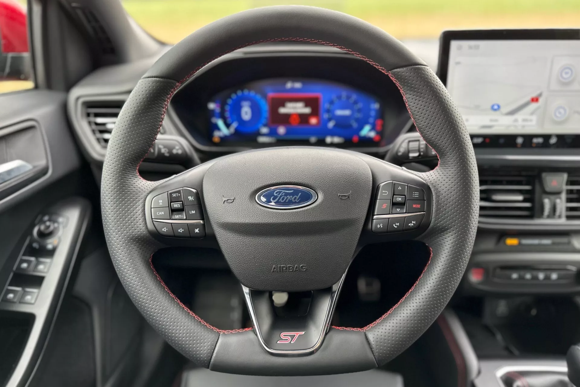 Vista interna della Ford Focus 2022 focalizzata sul volante sportivo rivestito in pelle con cuciture rosse e logo ST, cruscotto digitale e parte della console centrale con schermo dell'infotainment