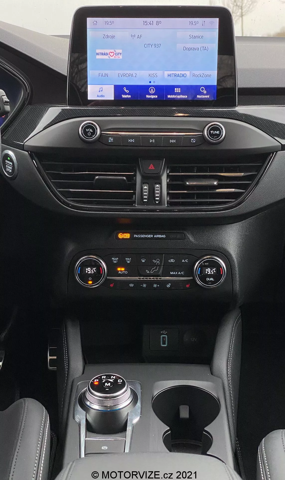 Vista interna della Ford Focus 2022 con enfasi sulla zona superiore centrale del cruscotto con il sistema di infotainment touchscreen prominente che mostra un'interfaccia colorata per media, connettività telefonica, navigazione e impostazioni dell'auto. L'immagine mostra anche il pannello di controllo del clima a doppia zona con pulsanti e manopole, così come i sbrinatori anteriori e posteriori. Sotto si trovano interruttori per assistenti alla guida come i sensori di parcheggio e il sistema start-stop, un selettore del cambio rotativo moderno, selettore della modalità di guida e pulsanti di controllo aggiuntivi, il tutto in un interno elegante e moderno con accenti in metallo spazzolato e materiali soft-touch.