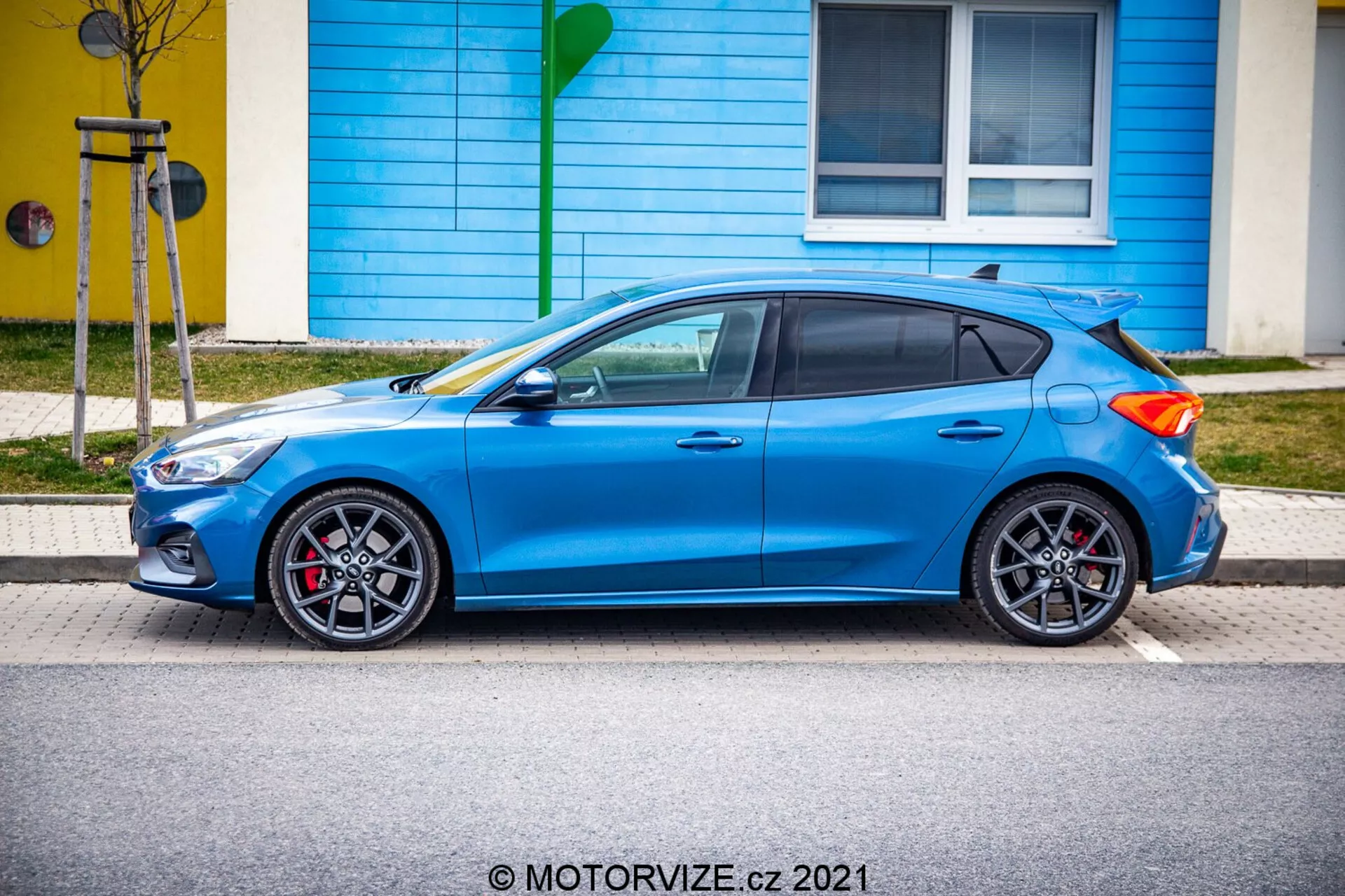 Vista di profilo laterale di una Ford Focus 2022 hatchback blu, che mostra il suo design sportivo con linee aerodinamiche eleganti, cerchi in lega con design a più razze e pinze dei freni rosse. Posizionata in parallelo al marciapiede, l'immagine cattura l'auto da un'angolazione leggermente elevata, fornendo una chiara vista degli specchietti retrovisori laterali nel colore della carrozzeria, dei finestrini oscurati e della piccola antenna nella parte posteriore del tetto. Sono visibili anche la portiera posteriore del hatchback e i paraurti anteriore e posteriore, insieme alle linee distintive che attraversano le porte. Lo spoiler posteriore e il tappo del carburante sul pannello posteriore evidenziano le caratteristiche pratiche e orientate alle prestazioni dell'auto.