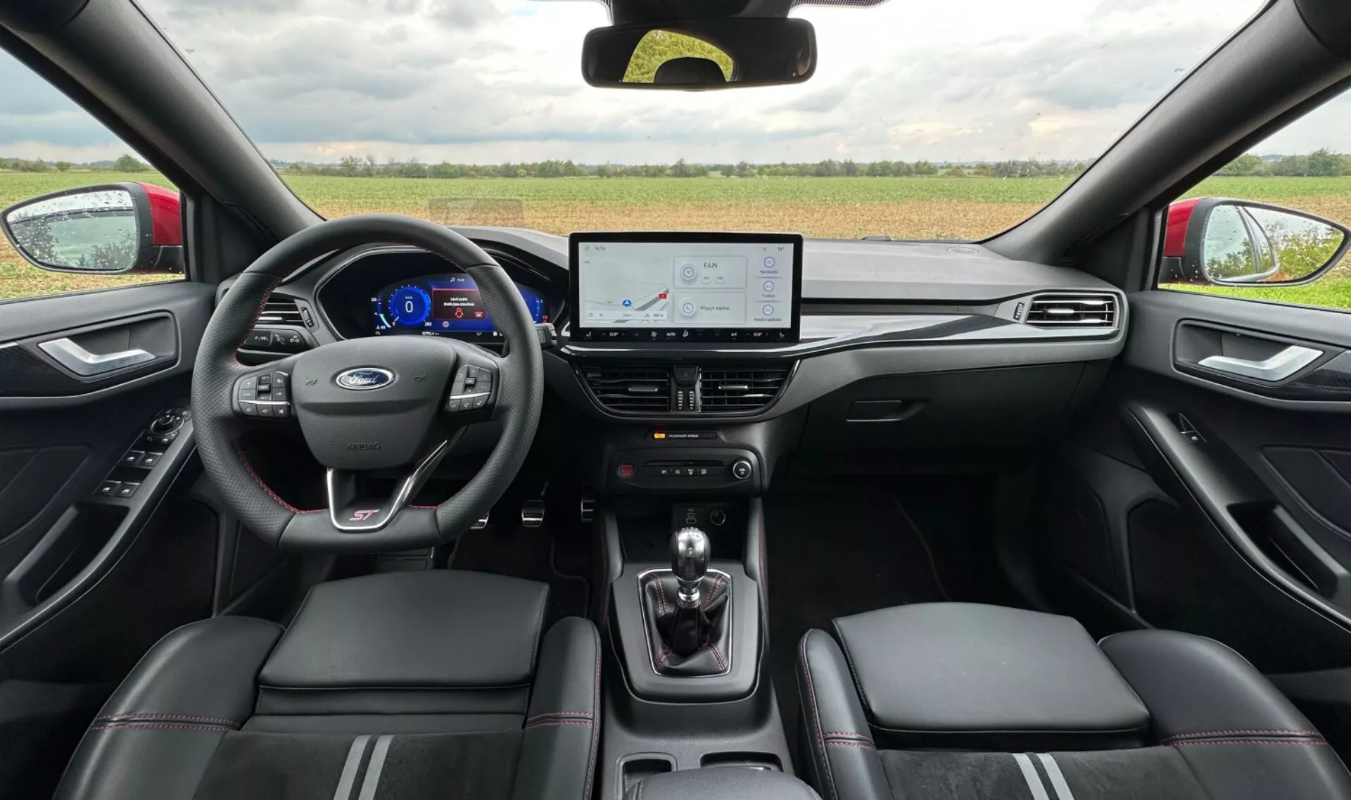 Vista interna del Ford Focus 2022 dal sedile posteriore che mostra cluster strumenti digitale con tachimetro blu, volante con logo Ford, schermo touchscreen infotainment con navigazione, selettore di trasmissione automatica e sedili in pelle con cuciture a contrasto