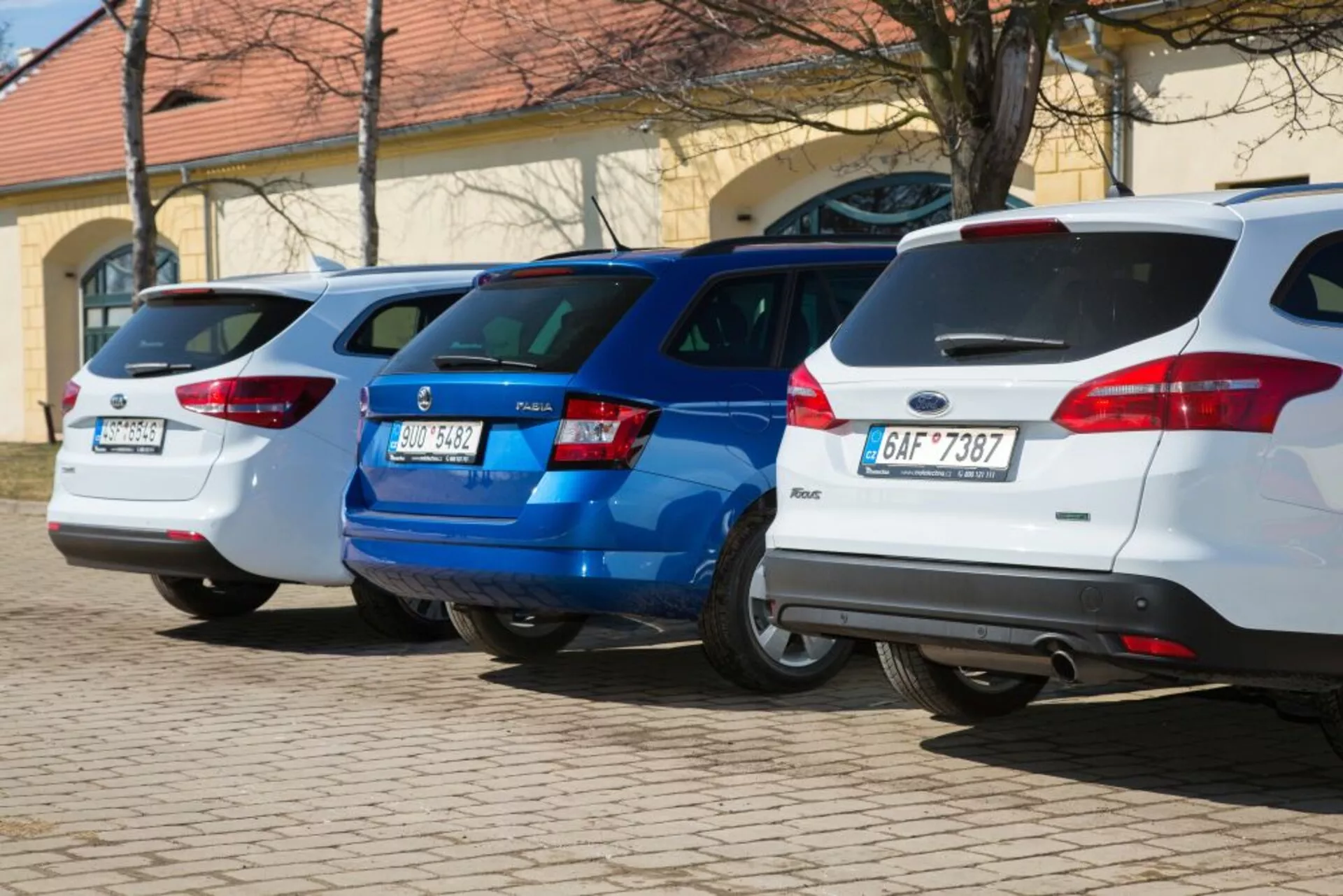 Una fila di tre hatchback, incluso un Ford Focus bianco del 2011, parcheggiati perpendicolarmente con i lati posteriori rivolti verso la fotocamera, mostrando le luci posteriori e le caratteristiche di design posteriore in un'area di parcheggio ben illuminata.
