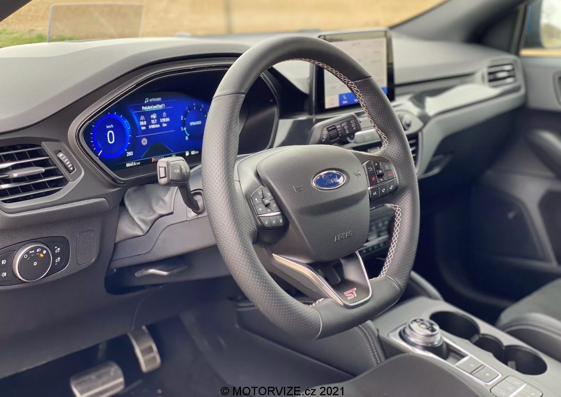 L'interno di una moderna Ford Focus 2022, mostrato dalla prospettiva del sedile del conducente, mette in risalto il volante con il logo Ford, rivestito in pelle con pulsanti di controllo, e un quadro strumenti completamente digitale che visualizza il tachimetro e altre funzioni. I materiali di alta qualità e le tecnologie avanzate, come il cambio automatico, i comandi dei fari e il sistema di infotainment centrale, indicano un design sofisticato con interfacce user-friendly.