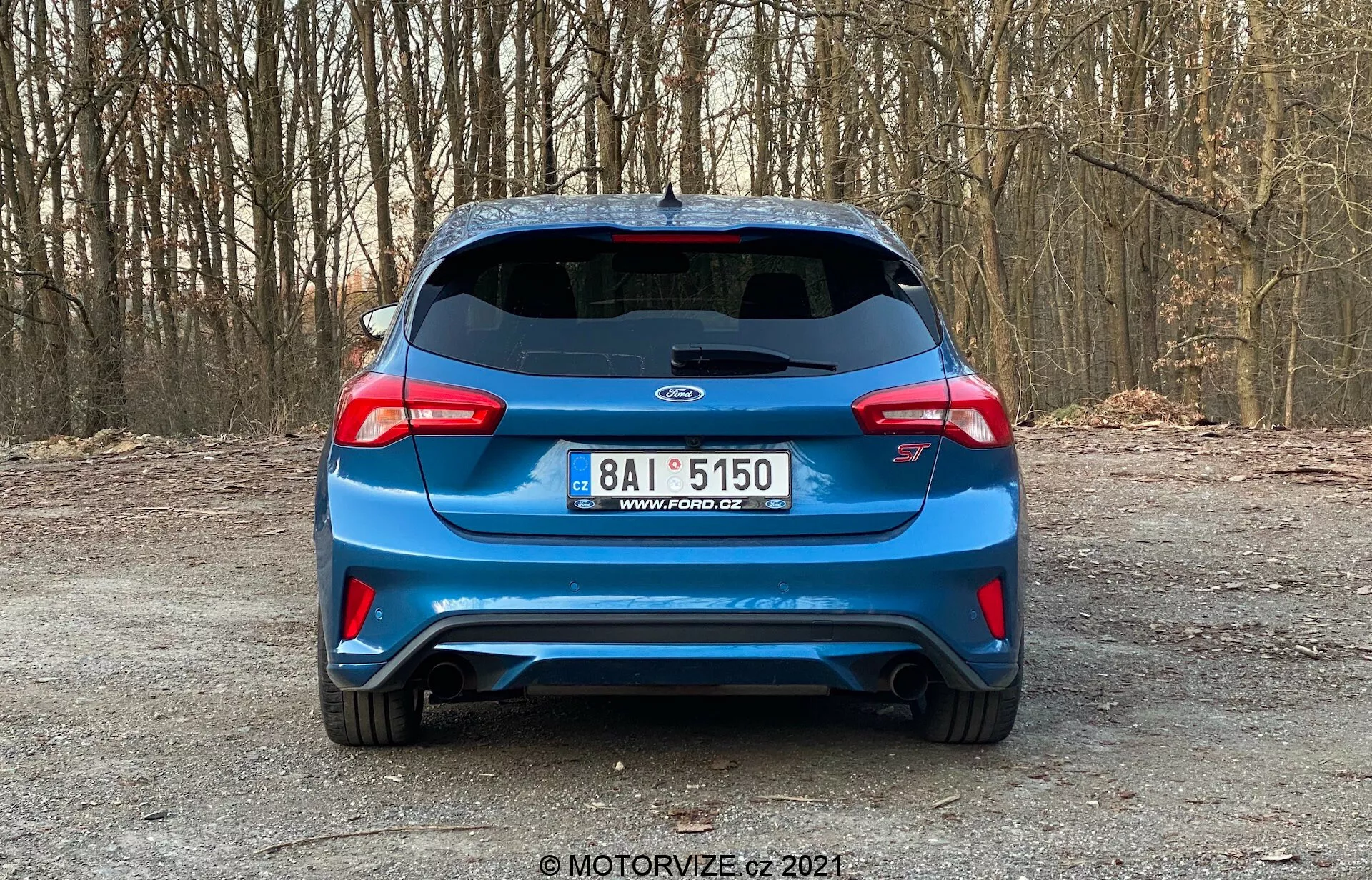 Vista posteriore di una Kia hatchback blu del 2022, probabilmente un modello Proceed GT, parcheggiata su una strada non asfaltata, possibilmente in una foresta. L'immagine mostra il cofano del bagagliaio, il paraurti posteriore e lo spazio tra i distintivi fanali posteriori avvolgenti. Le caratteristiche includono una targa in stile europeo centrata sotto il logo del produttore, l'emblema specifico del modello 'Proceed GT', uno spoiler montato sul tetto, tergicristallo posteriore, terza luce di stop, doppio scarico e un paraurti posteriore dallo stile aggressivo con elementi ventilati. La foto chiara e ben illuminata enfatizza il design sportivo del veicolo senza tecnologie avanzate visibili, scattata da un'angolazione perpendicolare standard.