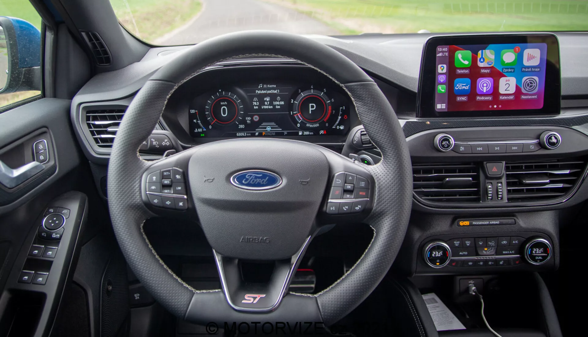 Vista interna della Ford Focus 2022 dalla prospettiva del conducente. I principali elementi includono un volante con logo Ford e logo sportivo 'ST', un cruscotto digitale che mostra velocità, selezione del cambio e stato della connettività. La console centrale ospita un sistema di infotainment con ampio schermo touch e integrazione per smartphone, controllo del clima a doppia zona e vari pulsanti di controllo.
