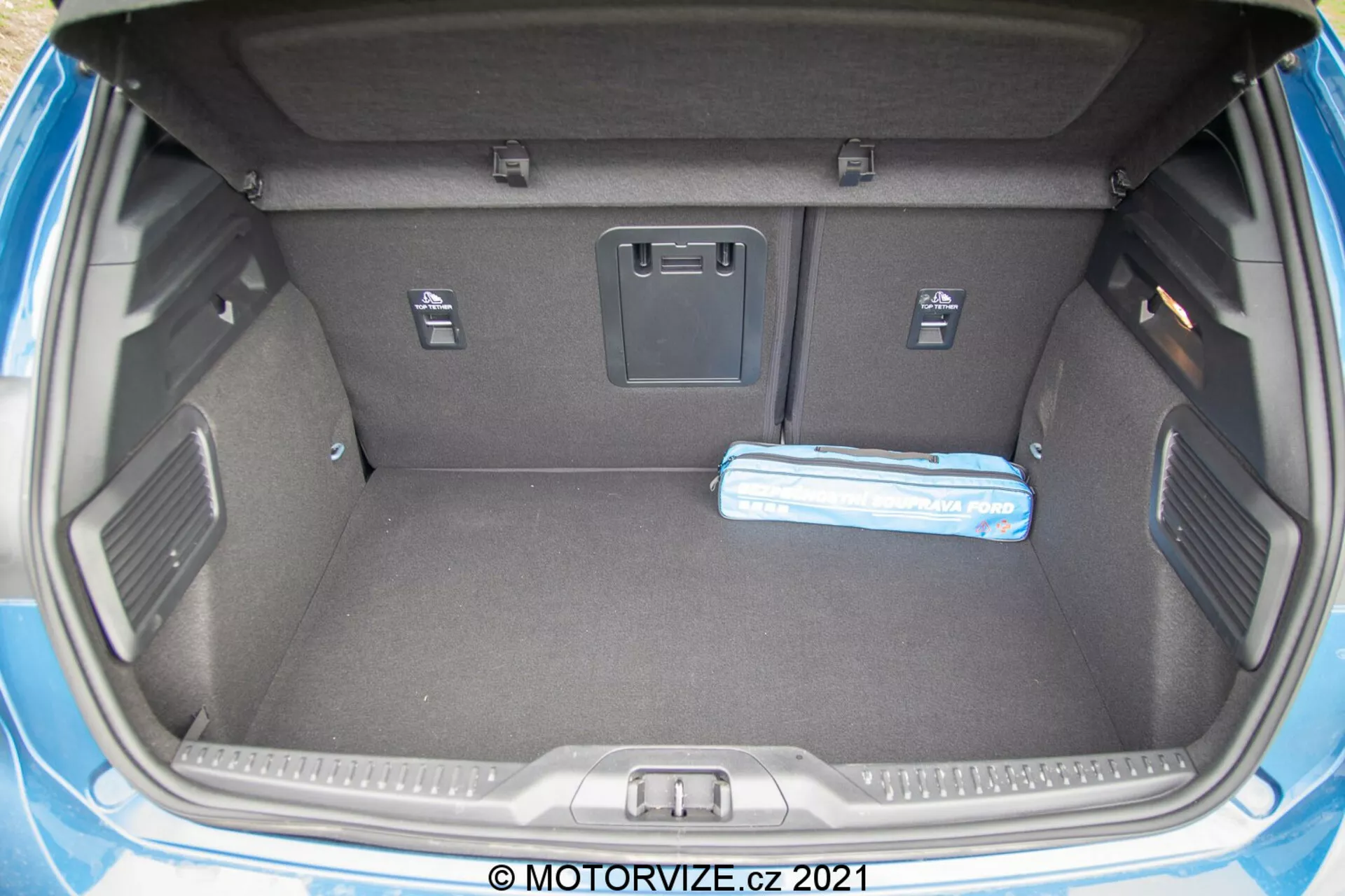 Immagine del bagagliaio di una Ford Focus 2022 da una prospettiva posteriore con il portellone aperto, che mostra un interno spazioso e rettangolare rivestito di moquette per ridurre il rumore e proteggere la superficie. Le caratteristiche includono un pavimento del bagagliaio piatto, rientranze per lo stoccaggio di piccoli oggetti, due ganci per fissare oggetti sciolti, piccoli scomparti per ulteriori spazi di stoccaggio, una presa di corrente a 12 volt, linguette per l'accesso ai sedili posteriori e una maniglia per chiudere il portellone. Un oggetto cilindrico blu, che sembra un ombrello piegato o una borsa portatile, è presente nel bagagliaio.