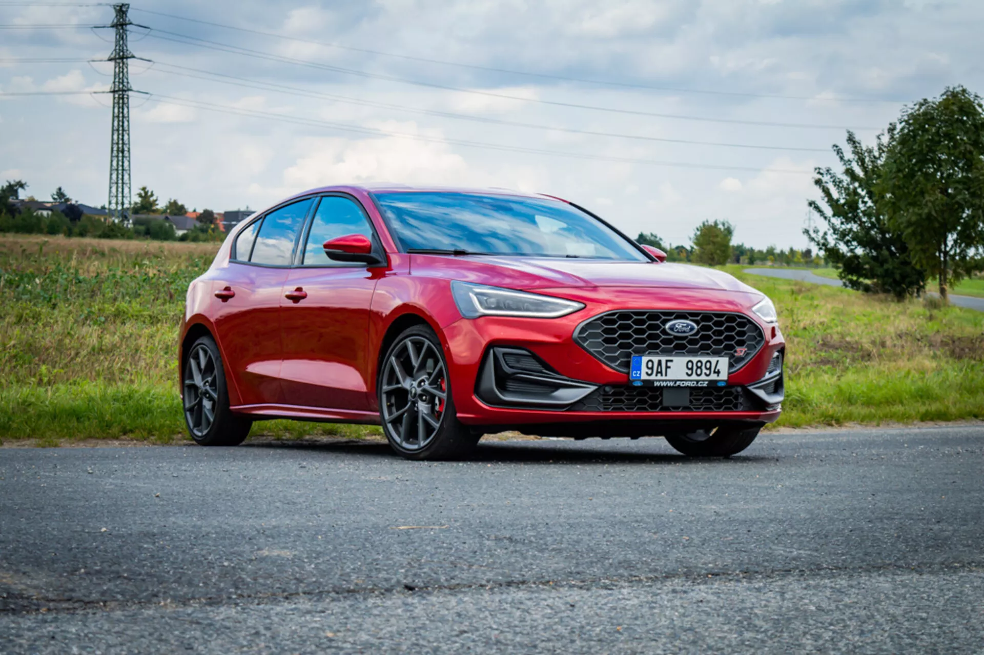 Sedan moderna Ford Focus 2022 parcheggiata su strada con cielo nuvoloso e paesaggio naturale sullo sfondo, evidenziando il suo colore rosso metallico, griglia anteriore sportiva con emblema, fari anteriori LED angolari, paraurti anteriore elegante e grandi ruote in lega a più razze da una prospettiva frontale tre quarti dal basso