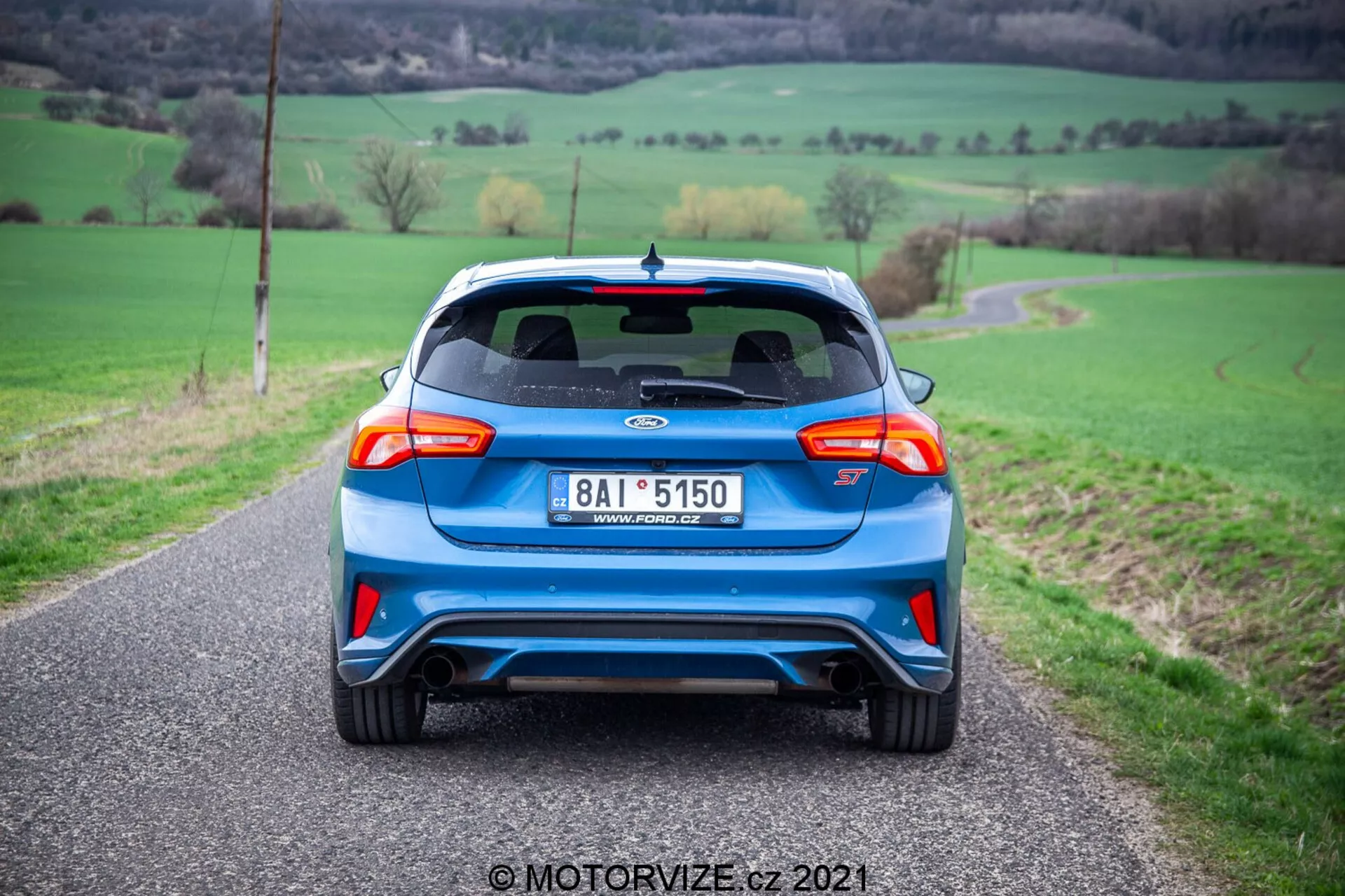Vista posteriore di un'auto blu con elementi di design moderni e sportivi, tra cui uno spoiler posteriore, luci posteriori eleganti, doppi tubi di scarico e un tergicristallo posteriore. Il veicolo si trova su una stretta strada rurale a due corsie con campi verdi su entrambi i lati.