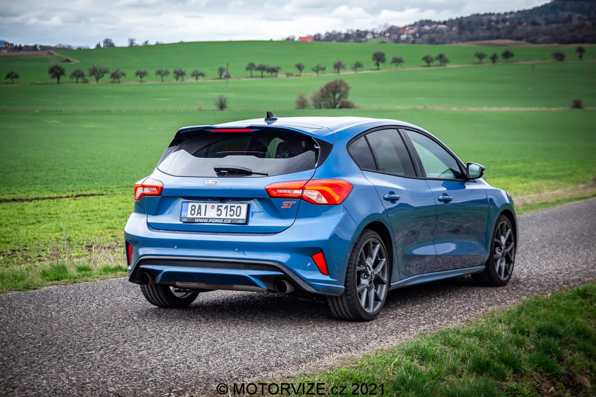 Vista tre quarti posteriore di una moderna berlina blu, posizionata su una strada stretta con un paesaggio verde sullo sfondo sotto un cielo nuvoloso. L'immagine cattura il lato destro e posteriore dell'auto, evidenziando il suo spoiler posteriore sportivo, il sistema di scarico doppio, i cerchi in lega a più razze e le luci posteriori distintive. L'aspetto del veicolo è enfatizzato dall'angolo basso, sottolineando il suo look dinamico. Sulla parte posteriore sono visibili anche un badge e una targa.