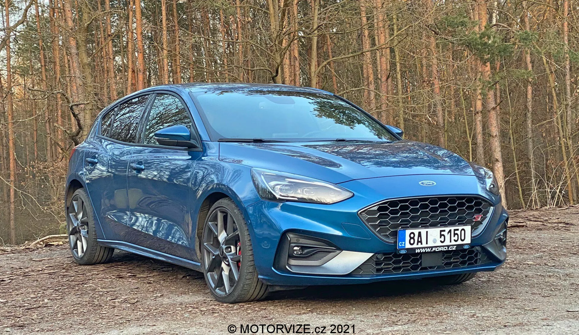 Ford Focus 2022 hatchback blu posizionato all'aperto in un ambiente autunnale avanzato o invernale. Catturato da un basso angolo anteriore di tre quarti, l'immagine enfatizza il design dinamico dell'auto, mostrando la grande griglia con motivo a nido d'ape, fari antinebbia integrati e fari a LED nel profilo anteriore. Il profilo laterale rivela cerchi in lega multirazza grandi, specchietti laterali in tinta con la carrozzeria con indicatori di direzione e finestre con bordi cromati. Una linea di carattere forte attraversa le maniglie delle porte. L'angolo decentrato fornisce profondità e prospettiva, evidenziando il battistrada e le condizioni degli pneumatici, con un ambiente ben illuminato per una chiara ispezione.