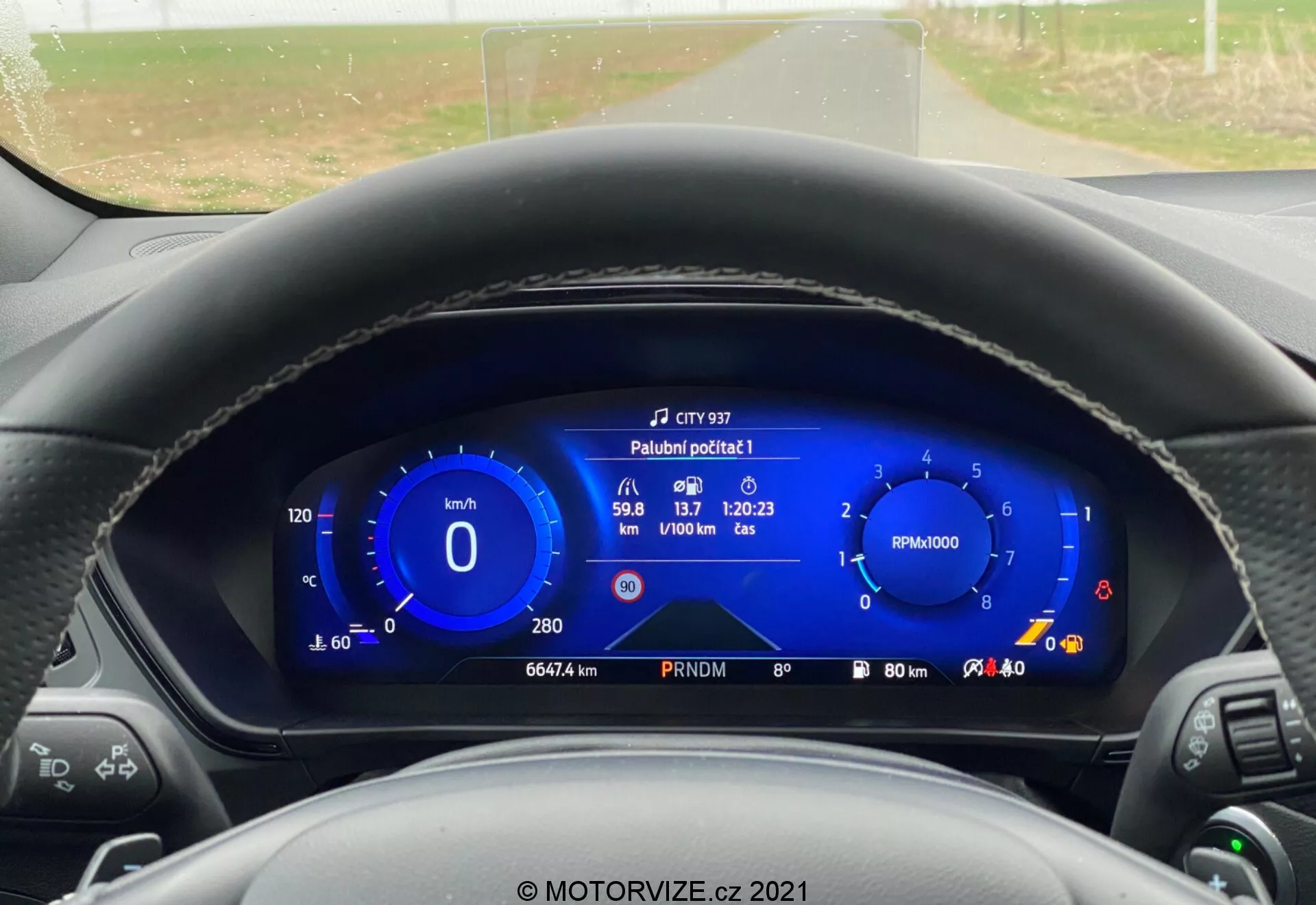 Vista in prima persona dal sedile del conducente di una Ford Focus 2022, che mostra un cruscotto completamente digitale. Il display digitale, evidenziato in un vibrante colore blu, include un tachimetro, un contagiri e varie informazioni sul veicolo come l'autonomia del carburante, la temperatura esterna, la velocità media, il tempo di viaggio e il consumo medio di carburante. Il volante è parzialmente visibile con le leve di controllo su entrambi i lati. Attraverso il parabrezza, si vede una strada rurale a due corsie, dritta, fiancheggiata da alberi e cespugli sotto un cielo coperto.