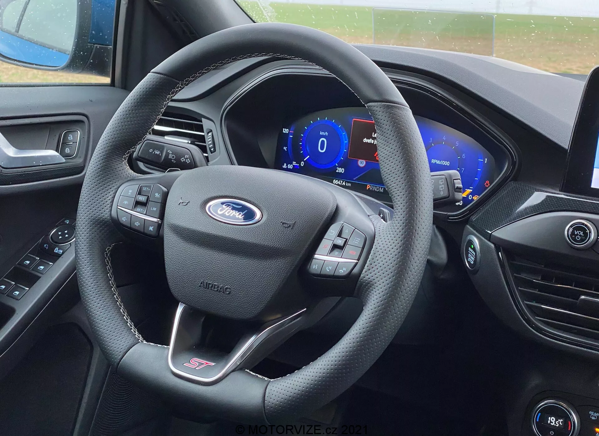 Chiara vista dell'interno del sedile del conducente in una Ford Focus 2022, che mostra il volante rivestito in pelle nera con il logo Ford prominente, badge della linea ST e controlli multifunzione. Dietro il volante c'è un quadro strumenti digitale che mostra 0 mph. Il cruscotto presenta contorni esteticamente piacevoli e uno schermo infotainment e prese d'aria parzialmente visibili.