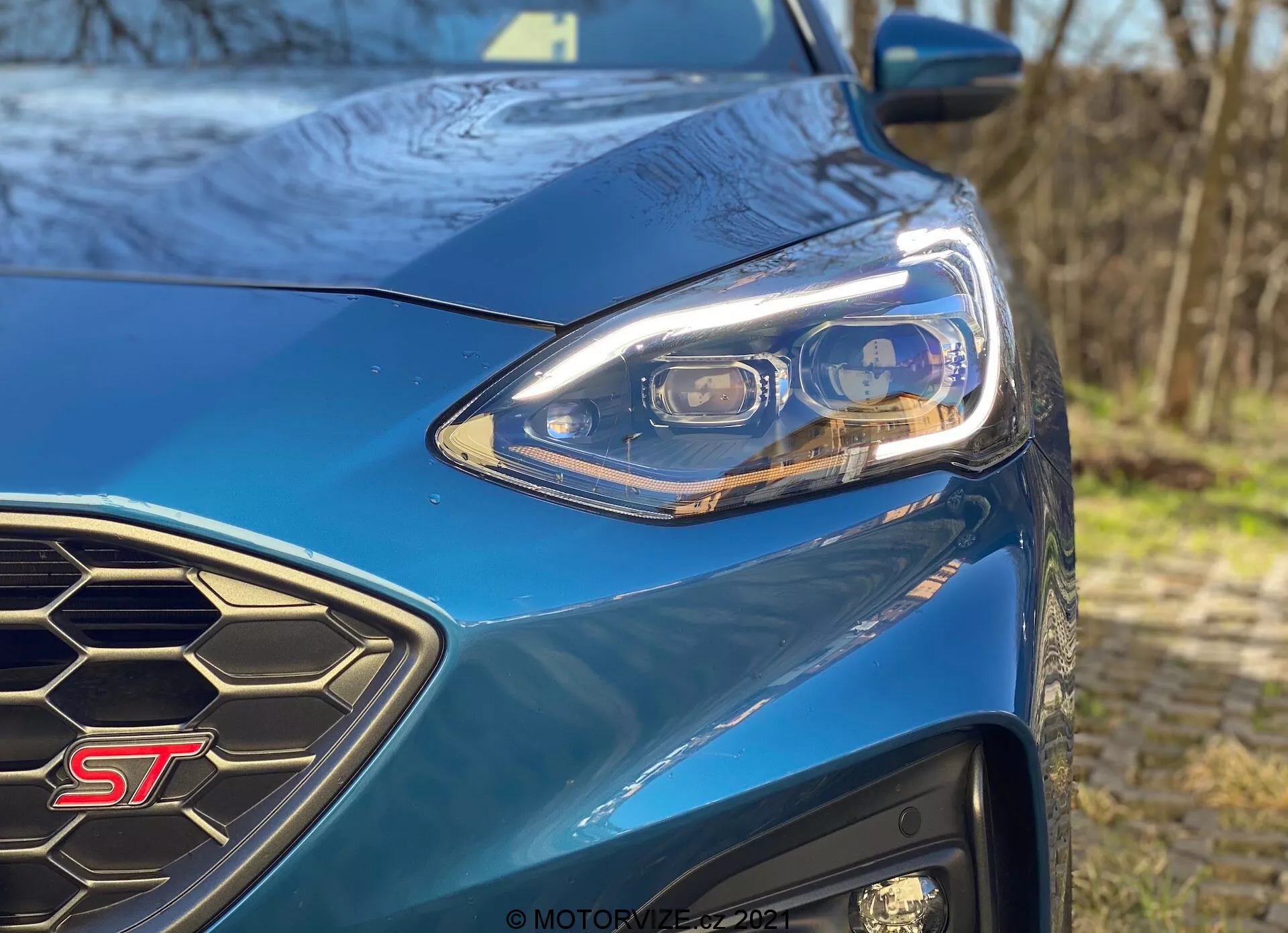 Primo piano del lato anteriore sinistro del modello ST blu della Ford Focus 2022, che mostra il design moderno dei fari a LED con molteplici elementi, la griglia a nido d'ape e il distintivo ST chiaramente visibile in un ambiente esterno molto illuminato.
