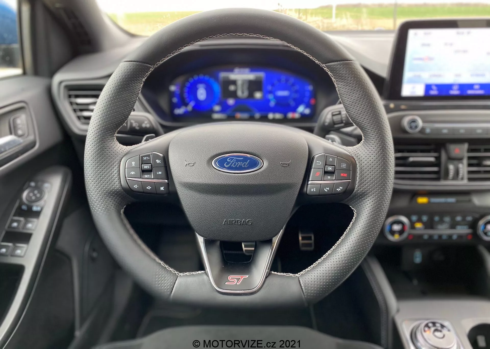Vista interna della Ford Focus ST 2022, che mostra il volante sportivo con cuciture in materiale simile alla pelle e pulsanti multifunzione per media e comandi vocali. Il cruscotto completamente digitale è illuminato, visualizzando tachimetro, contagiri e varie icone di assistenza elettronica. È visibile uno schermo centrale di infotainment, che indica funzionalità tecnologiche avanzate come la navigazione e i sistemi di connettività. L'immagine mette in evidenza l'interfaccia moderna e tecnologica del conducente dalla prospettiva del sedile del conducente, con la leva del cambio in 'Park'.