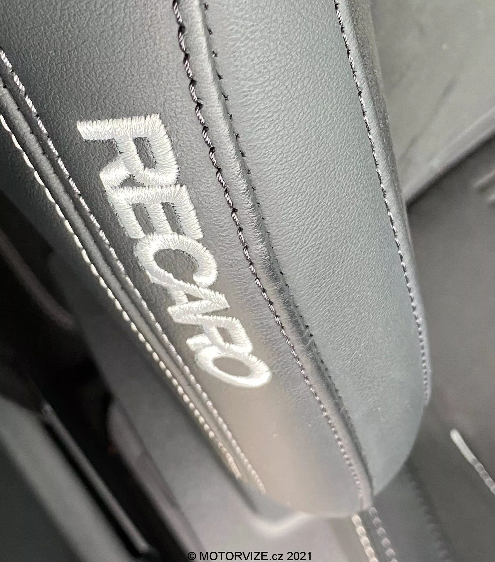 Vista ravvicinata del poggiatesta di un sedile dell'auto in una Ford Focus 2022, con ricamo 'RECARO' indicativo di sedili ad alte prestazioni. Il lato sinistro del poggiatesta è mostrato in modo prominente in una prospettiva inclinata dall'alto verso il basso, evidenziando il pregiato materiale in pelle o sintetico di qualità con cuciture dettagliate e doppie. L'illuminazione rivela la texture e la qualità del tessuto, enfatizzando il design ergonomico destinato a garantire comfort e supporto.