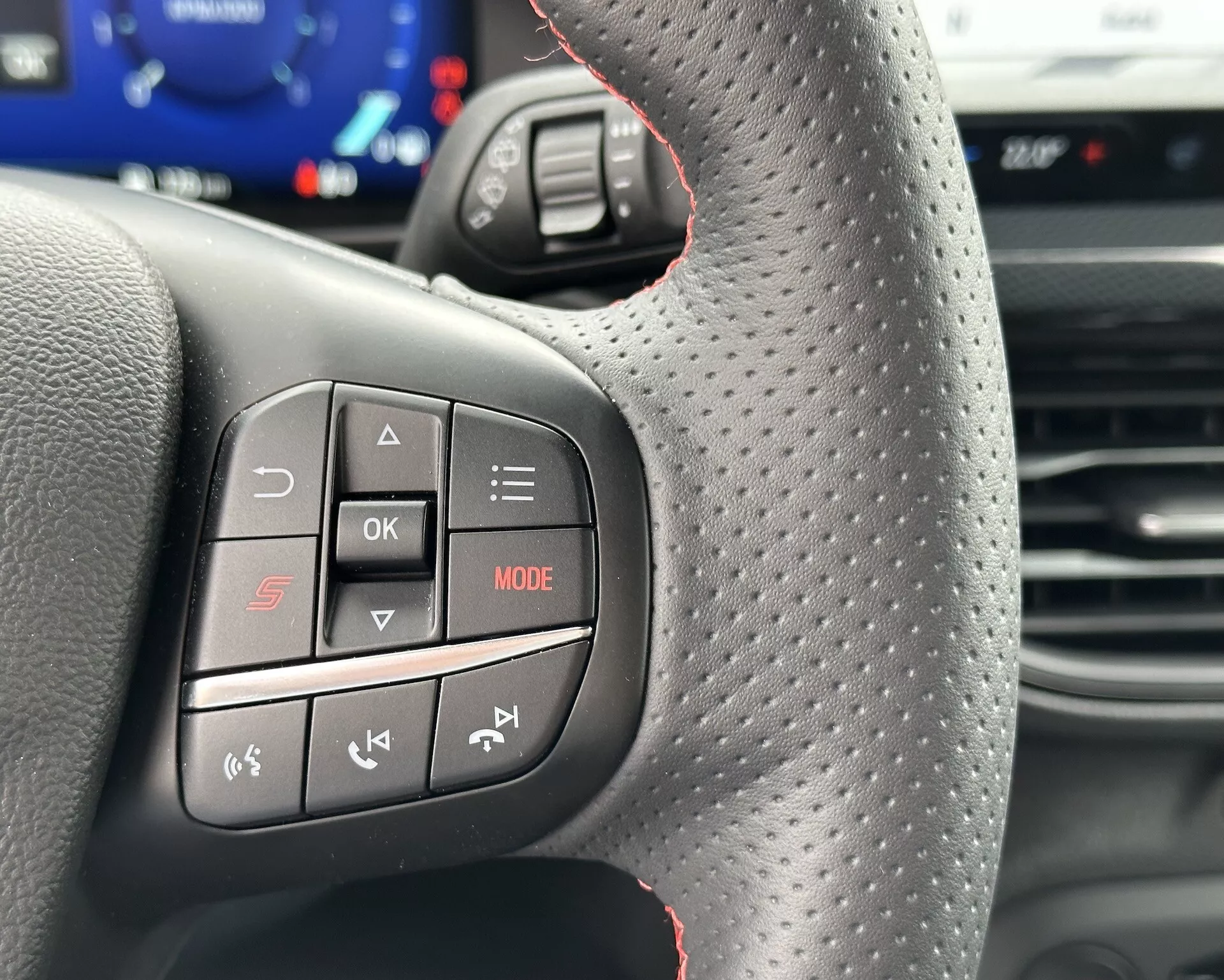 Vista ravvicinata del volante della Ford Focus 2022 che mette in mostra i controlli multifunzione dalla prospettiva del conducente, con pulsanti audio e telefono, cuciture rosse sportive e texture antiscivolo.