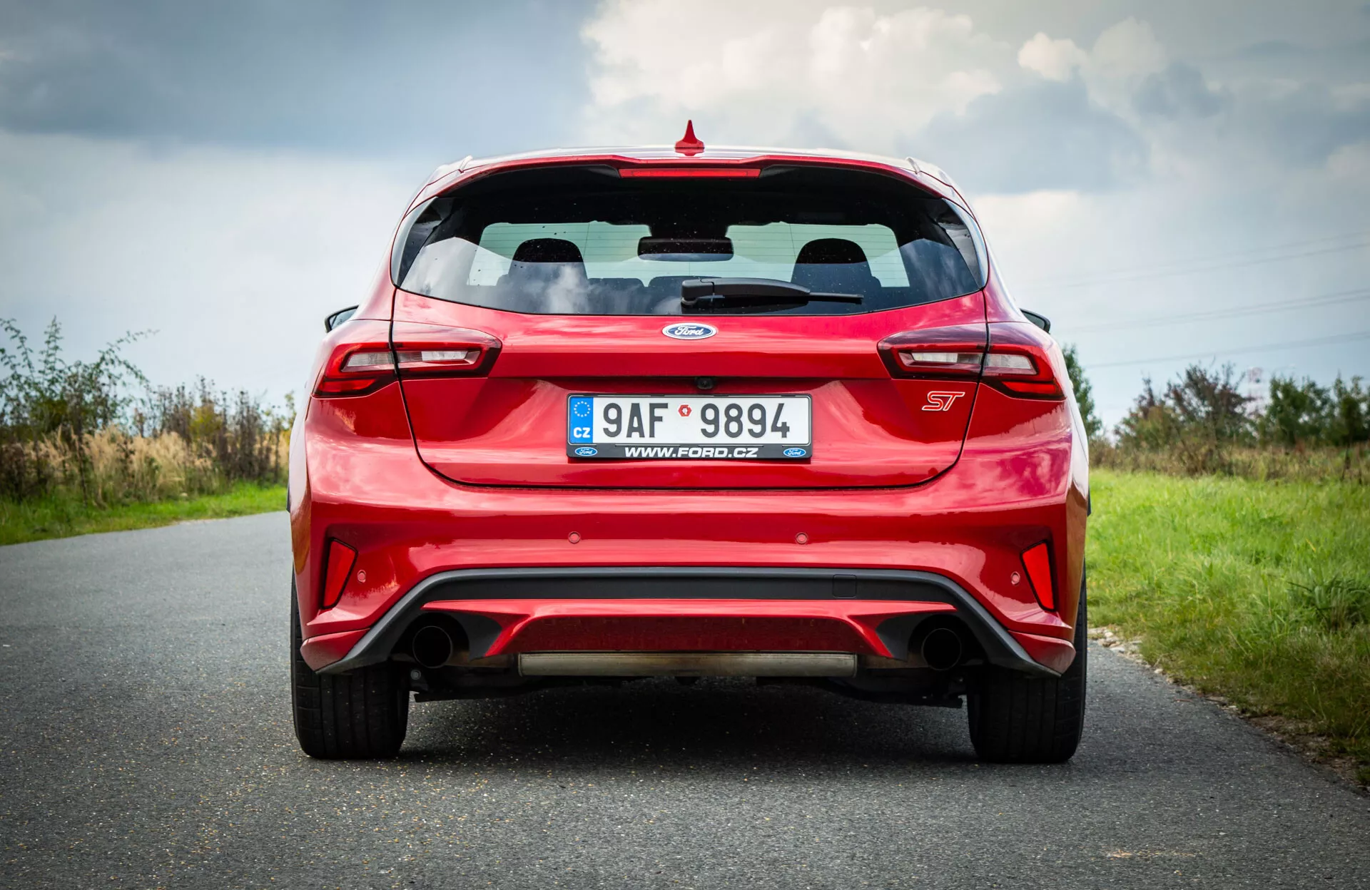 Ford Focus hatchback rosso 2022 catturato da una prospettiva posteriore e bassa che mostra la sua ampia postura, dotato di luci posteriori orizzontali che avvolgono i bordi, un finestrino posteriore chiaro con tergicristallo visibile, uno spoiler sportivo in cima, e il badge del modello dell'auto centrato sul portellone. Il retro mostra anche una targa, un paraurti sportivo con un elemento diffusore e prese d'aria, doppie punte di scarico integrate nel paraurti e luci riflettenti sui bordi del paraurti, impostato contro una strada e un paesaggio naturale sotto un cielo nuvoloso.