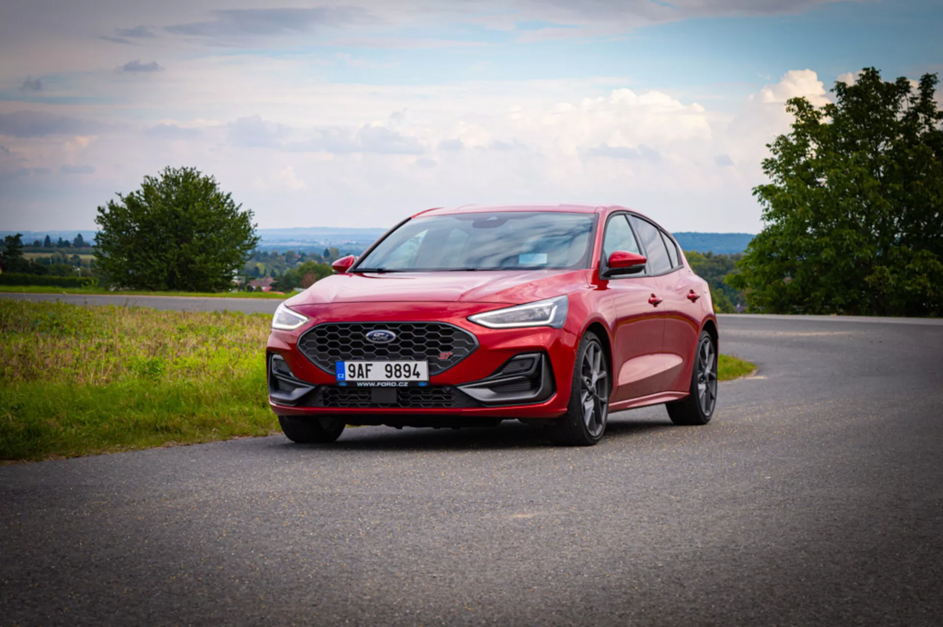 Ford Focus 2022 fotografata da un angolo basso frontale tre quarti che evidenzia la griglia anteriore con motivo a nido d'ape, fari LED snelli, cerchi in lega con design a più razze, cofano sagomato, specchietto laterale con indicatore di direzione integrato, carrozzeria rossa moderna con contorni aerodinamici, posizionata su una strada asfaltata con sfondo campestre