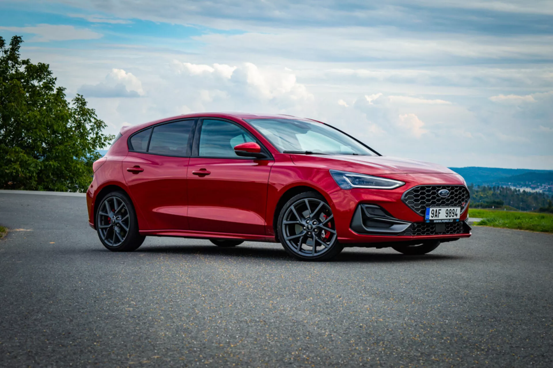 Vista frontale tre quarti di una Ford Focus hatchback rossa 2022 su una strada asfaltata con sfondo di campagna, evidenziando la sua griglia sportiva, fari LED, paraurti sagomato, ruote in lega e profilo dinamico.