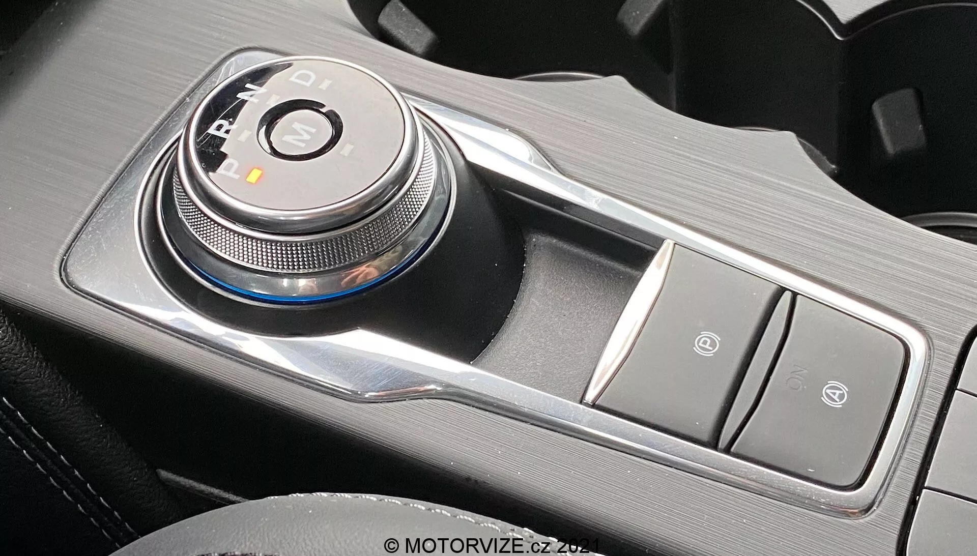 Immagine ravvicinata degli interni di una Ford Focus 2022 che mostra un avanzato selettore del cambio a rotella elettronico posizionato verso il lato del conducente sulla console centrale, nella posizione 'P' di parcheggio. Il selettore del cambio è circondato da indicatori LED per P, R, N, D, possibilmente S o B, riflettendo una trasmissione automatica con modalità sport o frenata. A destra, bottoni disposti verticalmente, probabilmente per il freno di stazionamento elettronico e la funzione Auto-Hold. La finitura metallica e i materiali testurizzati esaltano l'estetica premium e moderna alla luce naturale.