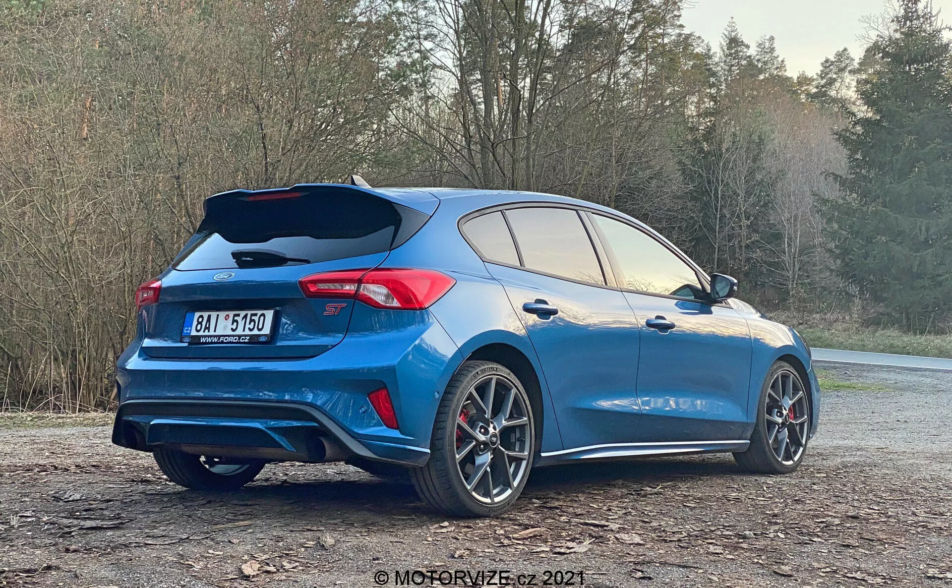 Vista posteriore a tre quarti di un'auto hatchback moderna, che mette in risalto i suoi elementi sportivi come uno spoiler sul tetto, modellazione aerodinamica e doppi tubi di scarico. L'auto è blu con dettagli neri o grigio scuro, grandi cerchi in lega multirazza con pinze dei freni visibili e luci posteriori a LED. La porta posteriore aperta rivela un ampio spazio di carico, suggerendo un facile accesso. Parcheggiata su un'area ghiaiosa con alberi sullo sfondo, questa immagine enfatizza il design del veicolo e le caratteristiche adatte a uno stile di vita attivo.