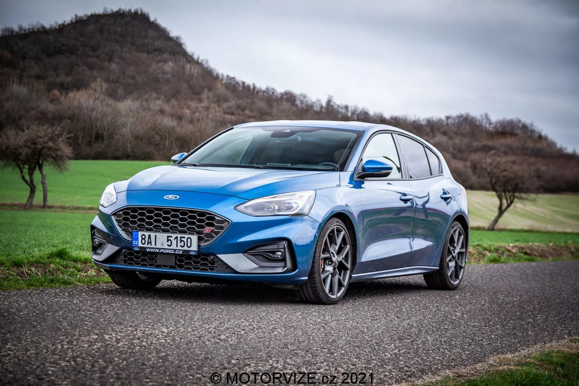 Ford Focus blu del 2022 catturato in una vista a tre quarti frontale da un'angolazione bassa, evidenziando il suo design aerodinamico, moderno e sportivo. Le caratteristiche evidenti includono la griglia a nido d'ape con un badge centrale, eleganti fari a LED, pieghe dinamiche sul cofano, grandi cerchi in lega a più razze, pneumatici a profilo ribassato, specchietti laterali con indicatori di direzione integrati e un indicatore laterale a lente chiara sul parafango anteriore. L'auto è a fuoco nitidamente su uno sfondo sfocato di un paesaggio collinare e un cielo nuvoloso, suggerendo che sia una berlina sportiva orientata alle prestazioni con caratteristiche avanzate di illuminazione automobilistica e sicurezza.
