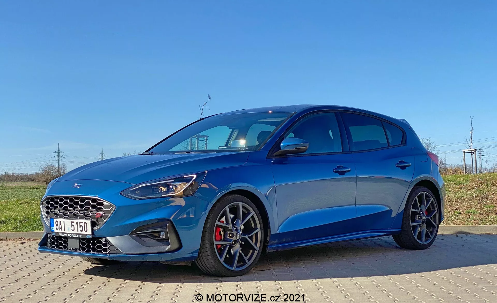 Immagine di una Ford Focus 2022 catturata da un angolo frontale di tre quarti, che mostra la griglia anteriore con motivo a nido d'ape, fari a LED, design moderno del paraurti con prese d'aria più grandi e un cofano elegante. Il veicolo hatchback blu metallizzato presenta una silhouette sportiva, pinze dei freni rosse, ruote multiraggio, linee di carattere affilate, passaruota sporgenti e specchietti laterali con indicatori di direzione integrati, il tutto evidenziato in condizioni di luce diurna chiara.