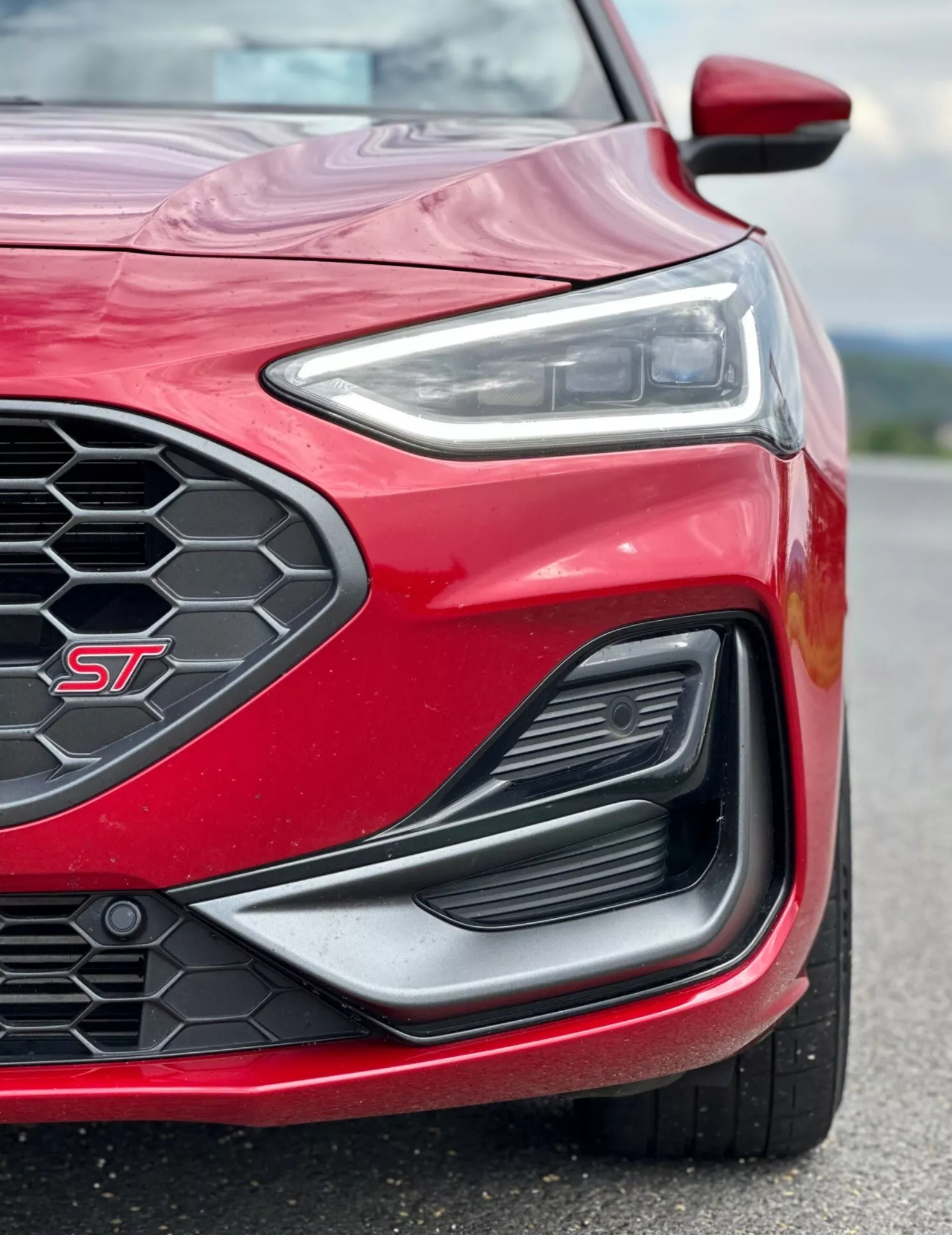 Vista ravvicinata dell'angolo anteriore sinistro di una Ford Focus ST 2022 in rosso, mostrando un design distintivo dei fari LED, griglia anteriore a nido d'ape, grande presa d'aria con trim aerodinamico e un'occhiata alla ruota e al parafango sportivi.