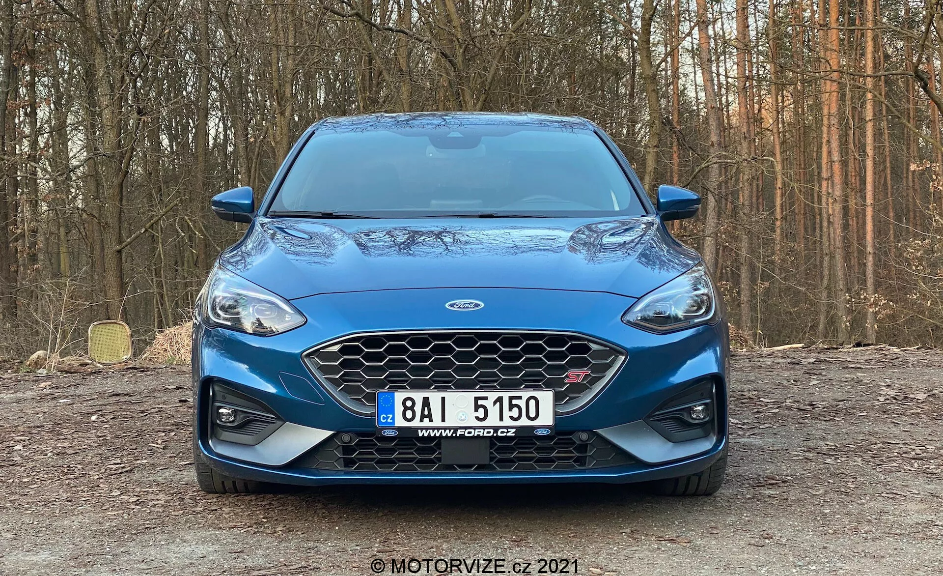 Vista frontale della Ford Focus 2022 catturata da un'angolazione leggermente bassa, che mostra la sua distintiva griglia a rete con il logo Ford centrale, eleganti fari a LED, fari fendinebbia e prese d'aria sul paraurti anteriore blu metallico. I contorni sportivi del cofano e i possibili sensori di parcheggio completano l'estetica moderna. Il veicolo è parcheggiato in un ambiente naturale, suggerendo un test drive o una sessione di revisione.