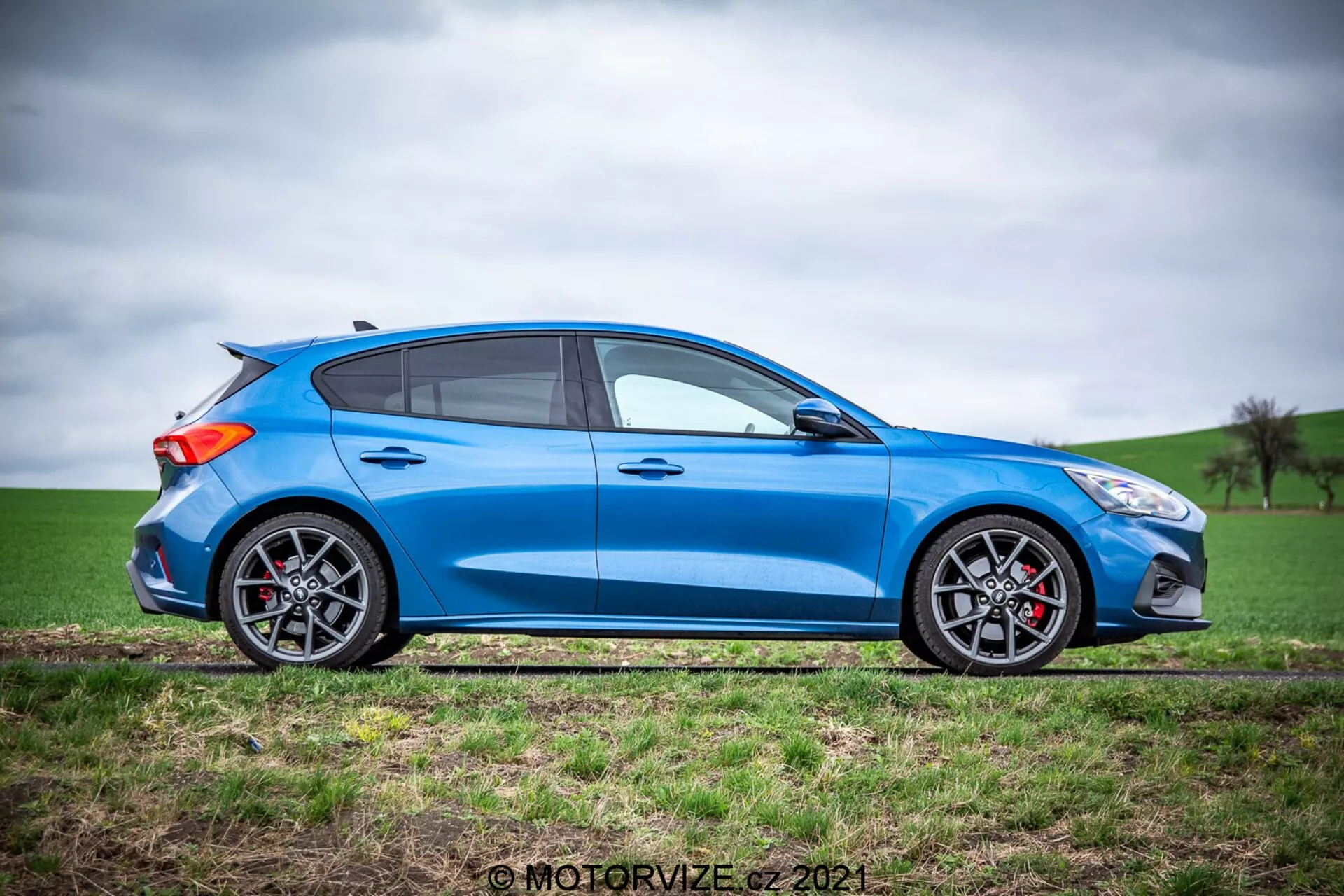 Vista laterale completa di un'elegante Ford Focus 2022 hatchback blu brillante alla luce naturale del giorno con una silhouette sportiva e aerodinamica, quattro porte, finestre con contorni neri e montante B nero, specchietti laterali neri, pinze dei freni rosse visibili attraverso ruote in lega con pneumatici a profilo basso e un piccolo spoiler posteriore su una superficie relativamente piatta adiacente a un campo con erba verde e un cielo coperto.
