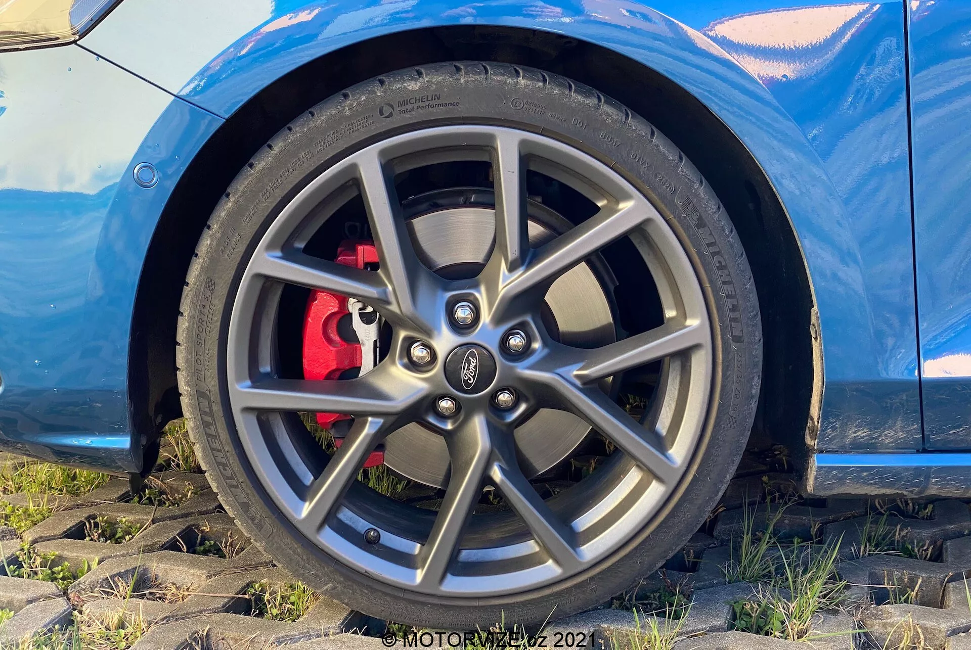 Vista ravvicinata del gruppo ruota e pneumatico della Ford Focus 2022 da una prospettiva bassa e leggermente angolata. L'immagine enfatizza il cerchio in lega multirazze, il pneumatico a basso profilo performante e la pinza del freno rossa dietro il cerchio, suggerendo un design sportivo. Nella parte superiore dell'immagine è visibile una porzione della carrozzeria dell'auto di colore blu metallico, contribuendo all'aspetto elegante e dinamico.