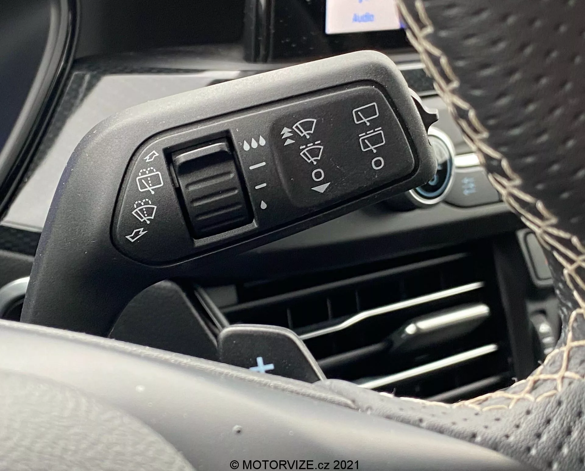 Vista ravvicinata della leva di controllo montata sulla colonna dello sterzo di una Ford Focus 2022 dalla prospettiva del conducente. L'immagine si concentra sulla leva di controllo sinistra con icone in rilievo per le impostazioni dei fari, gli indicatori di direzione e i tergicristalli. I riflessi brillanti sulla leva mettono in evidenza la qualità del materiale. Sullo sfondo sfocato si possono distinguere le prese d'aria e gli elementi del cruscotto.