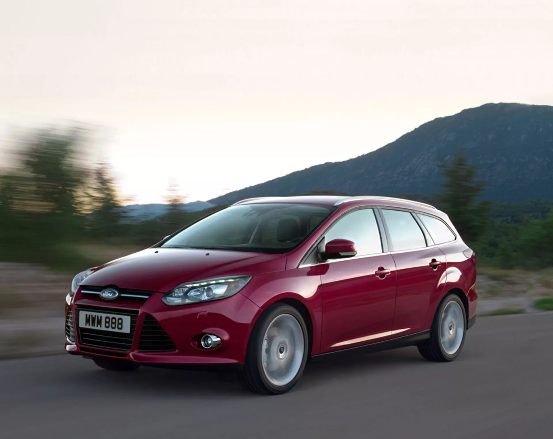 Station wagon Ford Focus rosso 2011 in movimento su una strada d'asfalto, che mostra il suo design moderno, griglia anteriore prominente con logo Ford, fari inclinati all'indietro, linee scolpite sul cofano e ruote in lega, sotto condizioni di illuminazione serale