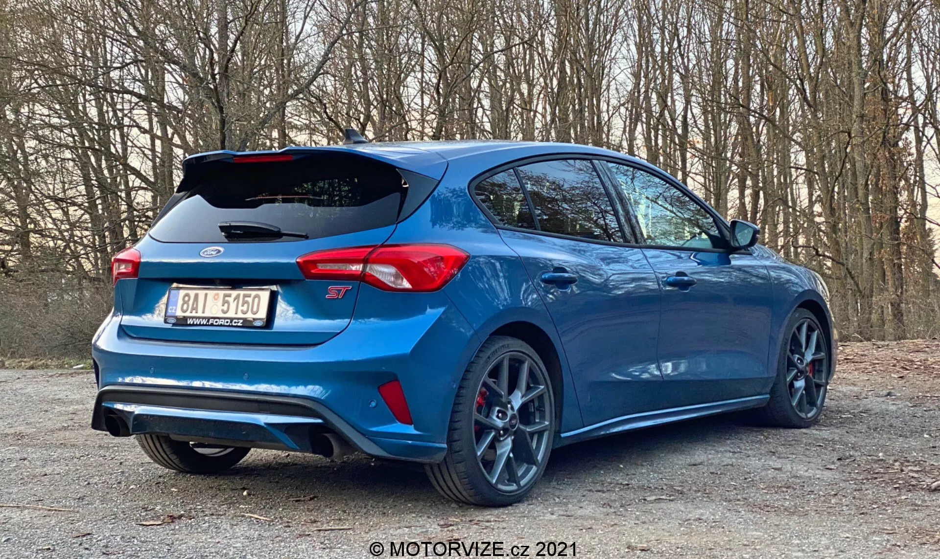 Un hatchback moderno Ford Focus 2022 parcheggiato in un'area appartata della strada con alberi sullo sfondo, visto da un angolo basso di tre quarti posteriore. Sono visibili le caratteristiche posteriori dell'auto, il profilo laterale, le ruote, lo spoiler aerodinamico, i doppi scarichi, il diffusore posteriore, le luci posteriori a LED, il distintivo del modello, il paraurti posteriore distintivo, i cerchi sportivi a più raggi con pneumatici a basso profilo, la pinza del freno rossa ad alte prestazioni, la vernice metallica blu e i vetri oscurati, evidenziando elementi di design automobilistico, prestazioni e tecnologia di sicurezza.
