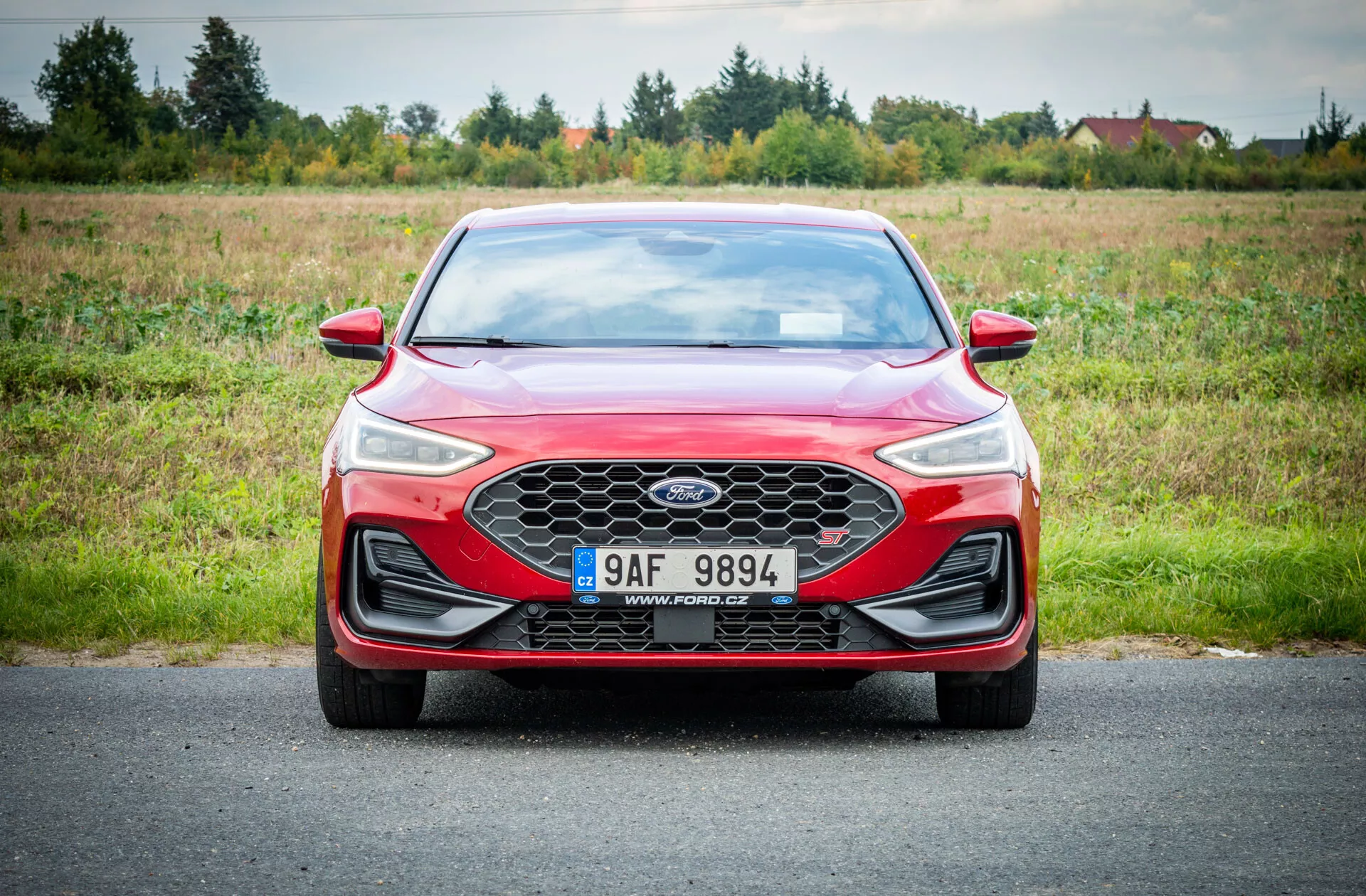 Sedan rosso Ford Focus 2022 su strada con sfondo naturale erboso sotto illuminazione nuvolosa che mostra vista frontale con griglia a nido d'ape, fari LED e design aerodinamico